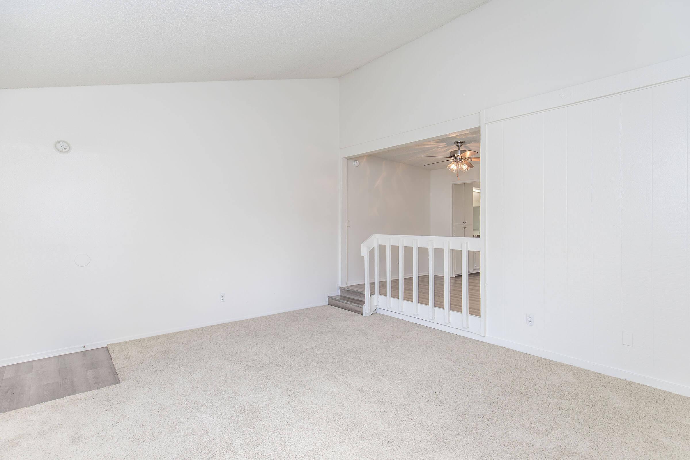 A spacious, empty living room with light-colored walls and a carpeted floor. There is a partial wall with a railing leading to another room, which is visible through an opening. The room features a ceiling fan and is well-lit, creating a bright and airy atmosphere.