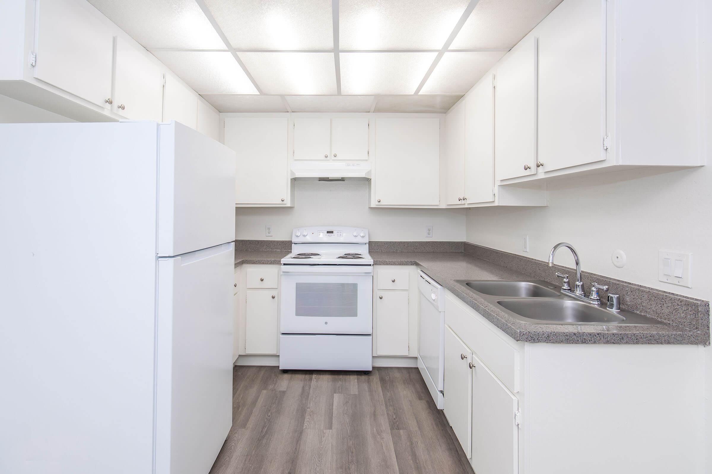 A modern kitchen featuring white cabinets, a white refrigerator, a white stove with an oven, and a double sink. The countertops are a light gray, and the flooring is a wood-like laminate. The ceiling has fluorescent lighting, providing bright illumination in the space.