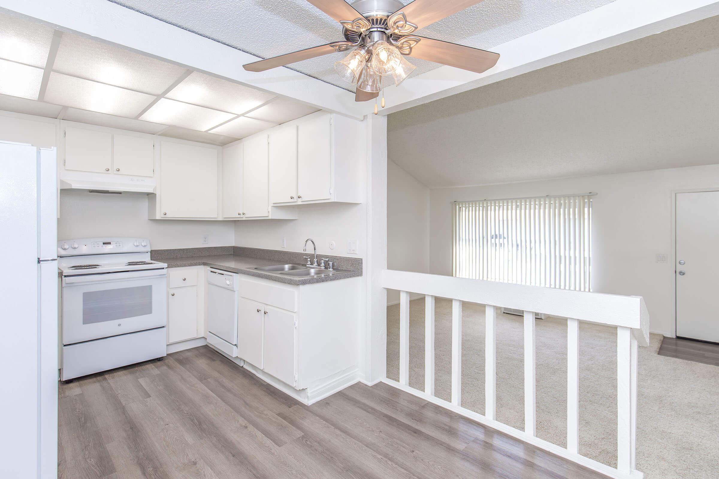 A bright, modern kitchen with white cabinetry and appliances, featuring a sink and a ceiling fan. The kitchen connects to a spacious living area with beige carpet and large vertical blinds on a window. The layout suggests an open floor plan, enhancing the feeling of space.