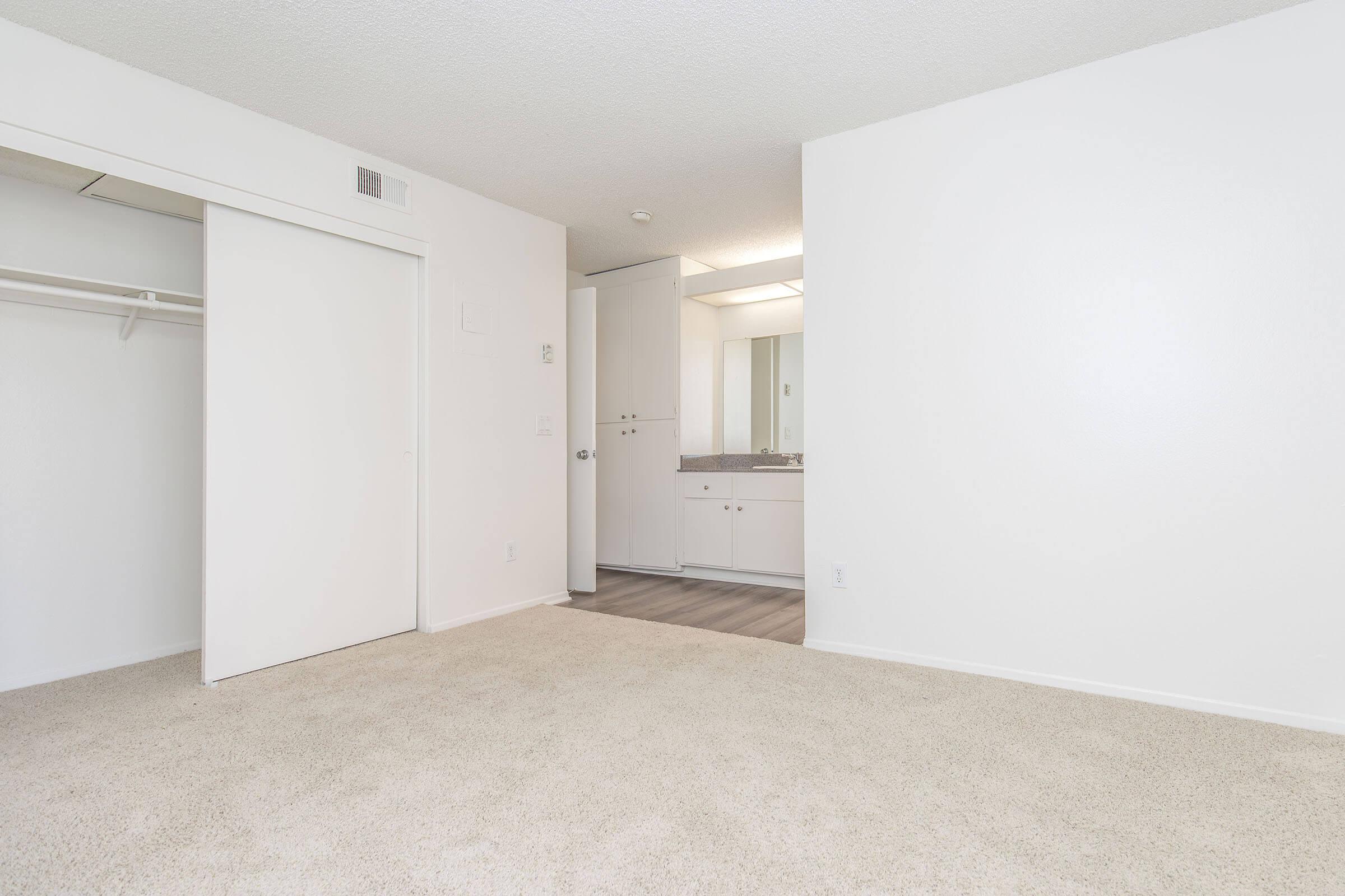 An empty, well-lit room featuring beige carpet and white walls. On one side, there's a closet with sliding doors, and on the opposite side, a small vanity area with a mirror and a sink. The flooring transitions to a different surface leading to a bathroom area.