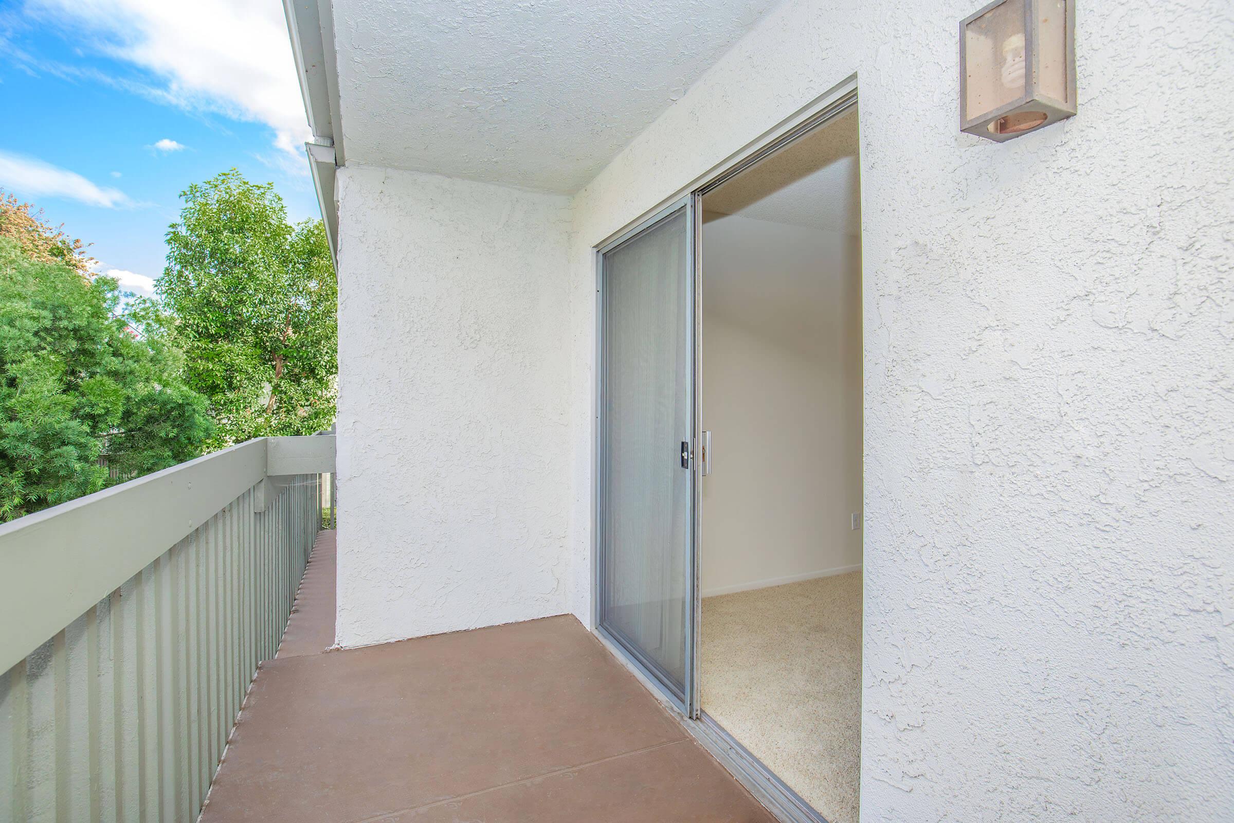 A view of a balcony with a sliding glass door leading to an interior space. The exterior walls are painted white, and there is a railing along the edge. Lush green trees are visible in the background, under a clear blue sky. The floor is a neutral color, and the interior appears well-lit.