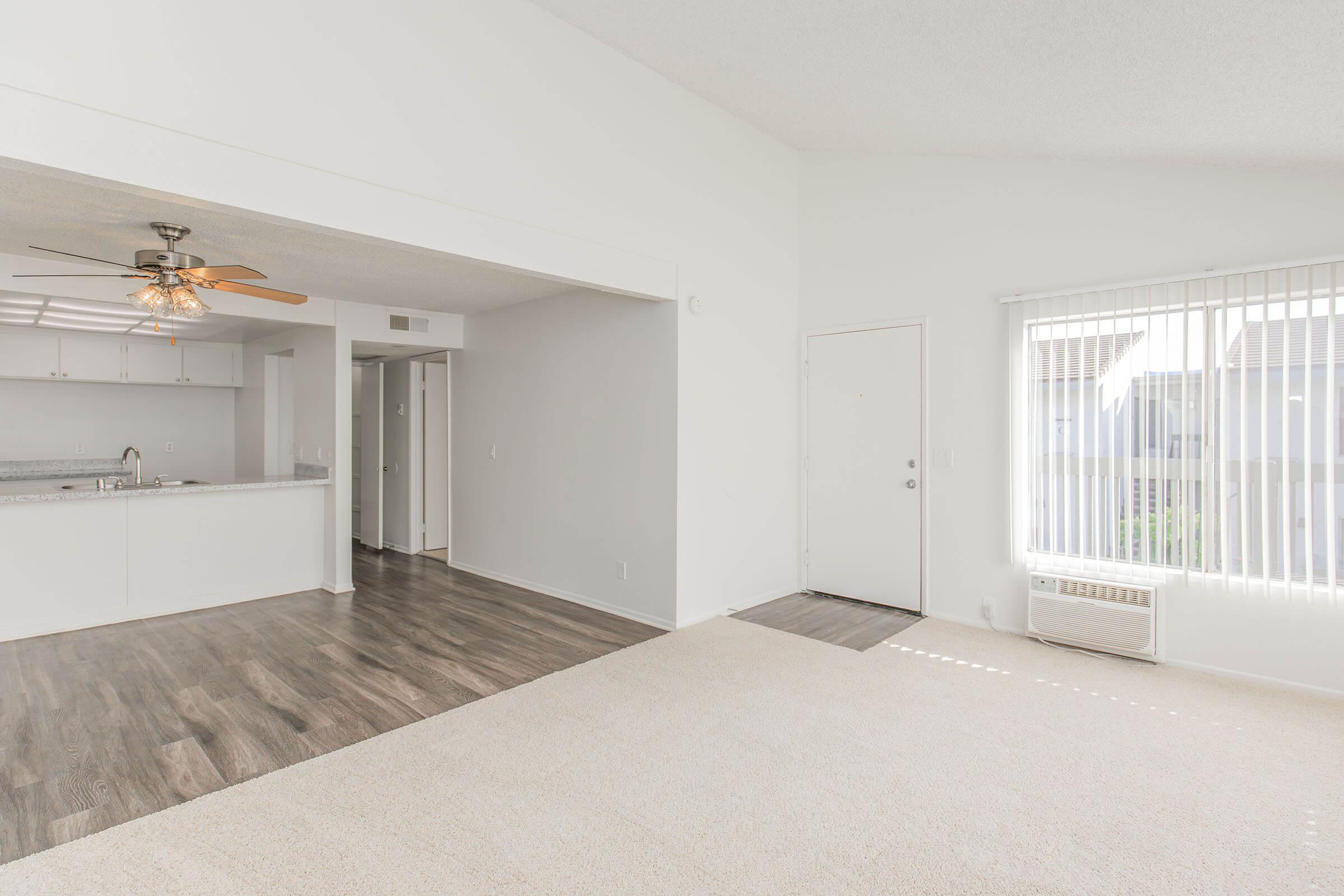A brightly lit, spacious living area featuring light-colored walls and flooring. The room has a ceiling fan and an air conditioning unit. A doorway leads to a kitchen with white cabinetry visible in the background. Natural light streams through a window with vertical blinds, and the floor is carpeted in a soft beige.