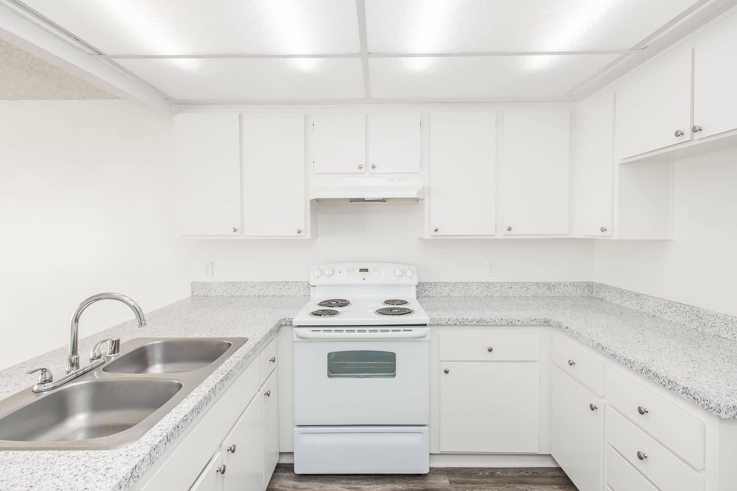 A modern kitchen featuring white cabinetry, a gray speckled countertop, and a white stove with four burners. The kitchen has a double sink, overhead cabinets, and bright lighting, creating a clean and spacious appearance.