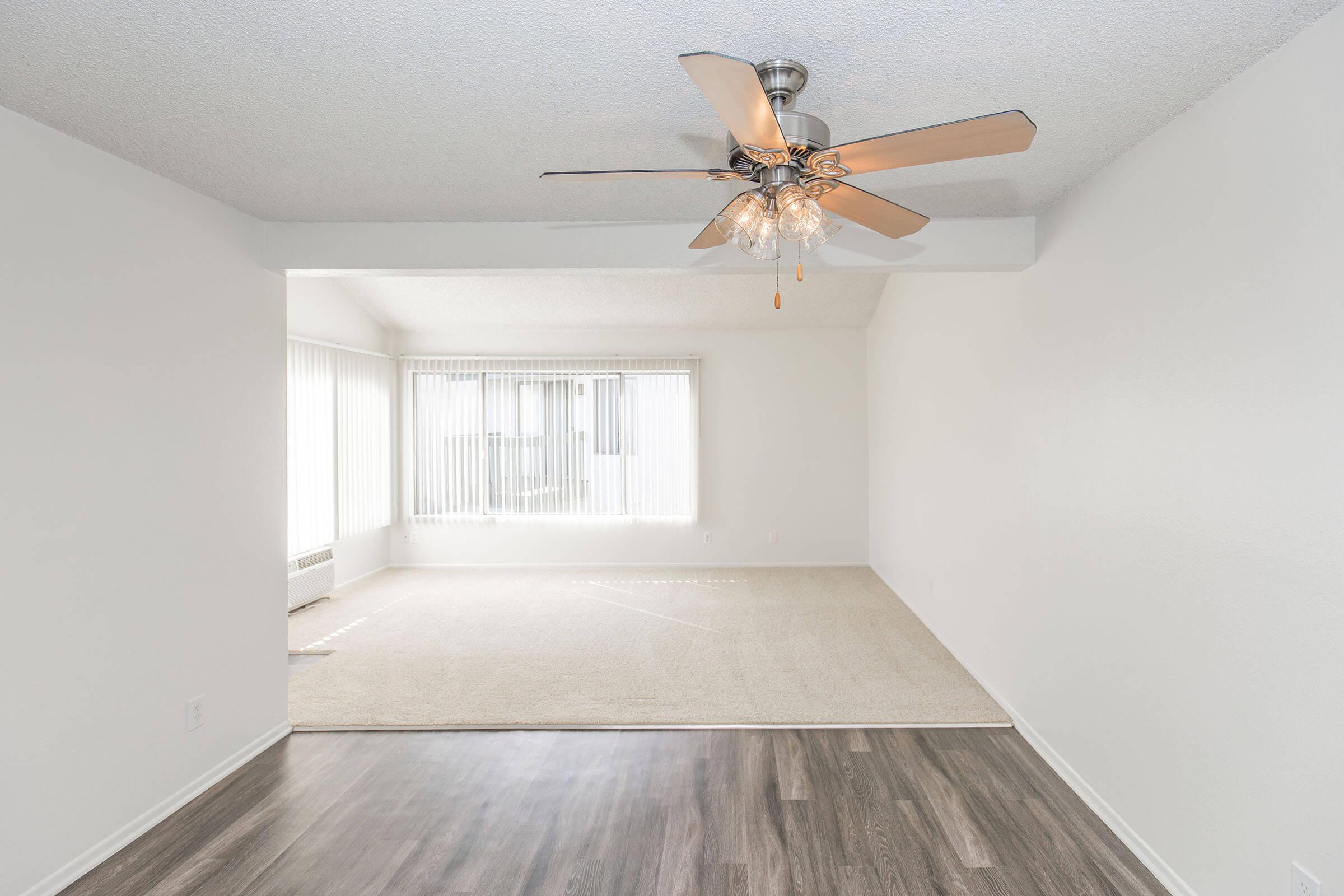A spacious, well-lit living area featuring a ceiling fan with wooden blades. The room has light-colored walls and large windows with vertical blinds, allowing natural light to fill the space. The flooring is a mix of carpet and hardwood, creating a cozy yet modern atmosphere.