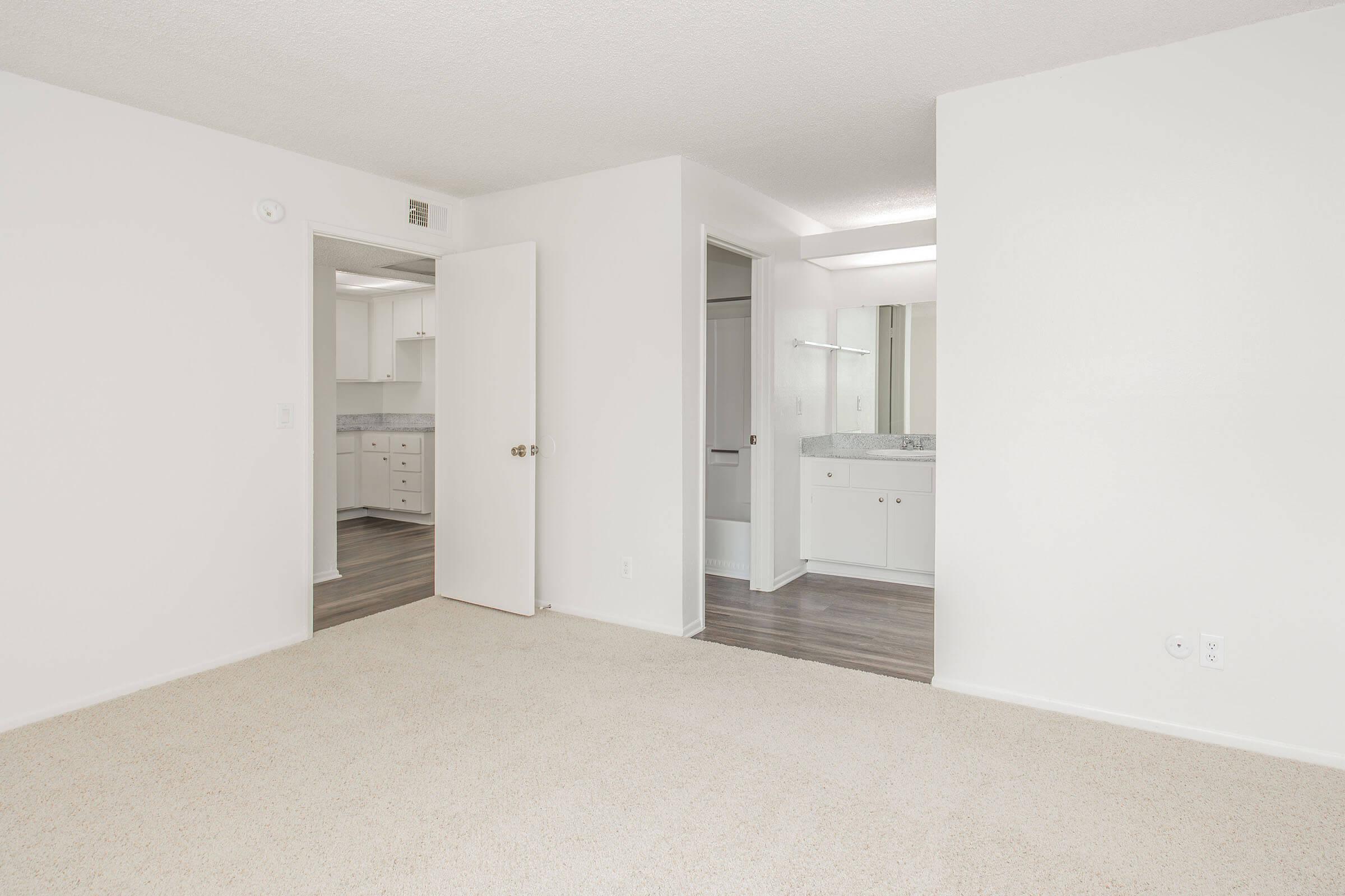 A spacious, well-lit room with light-colored walls and carpet. It features an open layout leading to a kitchenette and bathroom area. The doorways are visible, indicating access to other rooms, and the overall atmosphere is clean and modern.