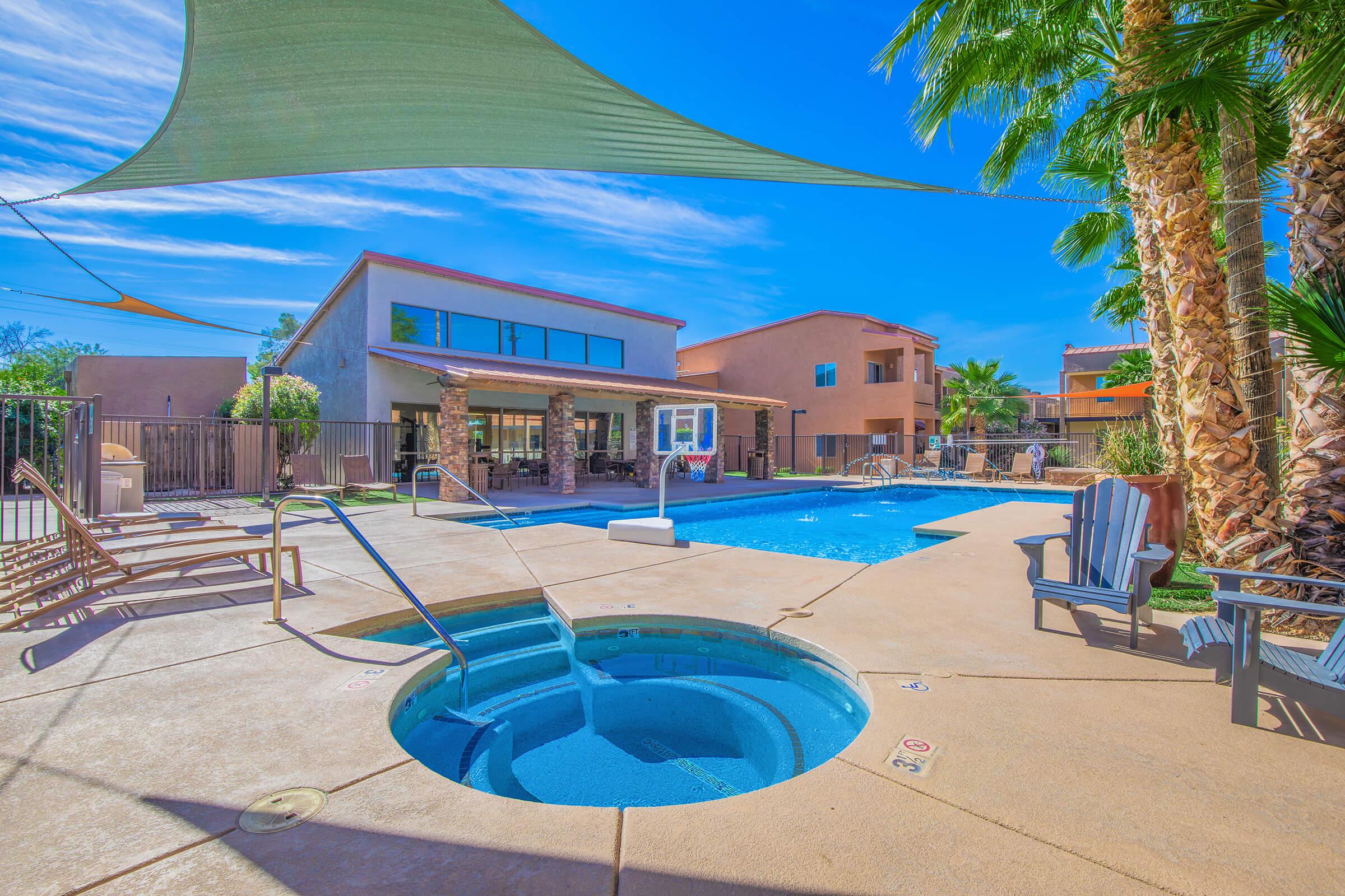 A sunny outdoor swimming pool area featuring a large pool, a hot tub, and lounge chairs. There are palm trees and shade sails nearby, along with a colorful building in the background. The setting is inviting and perfect for relaxation.