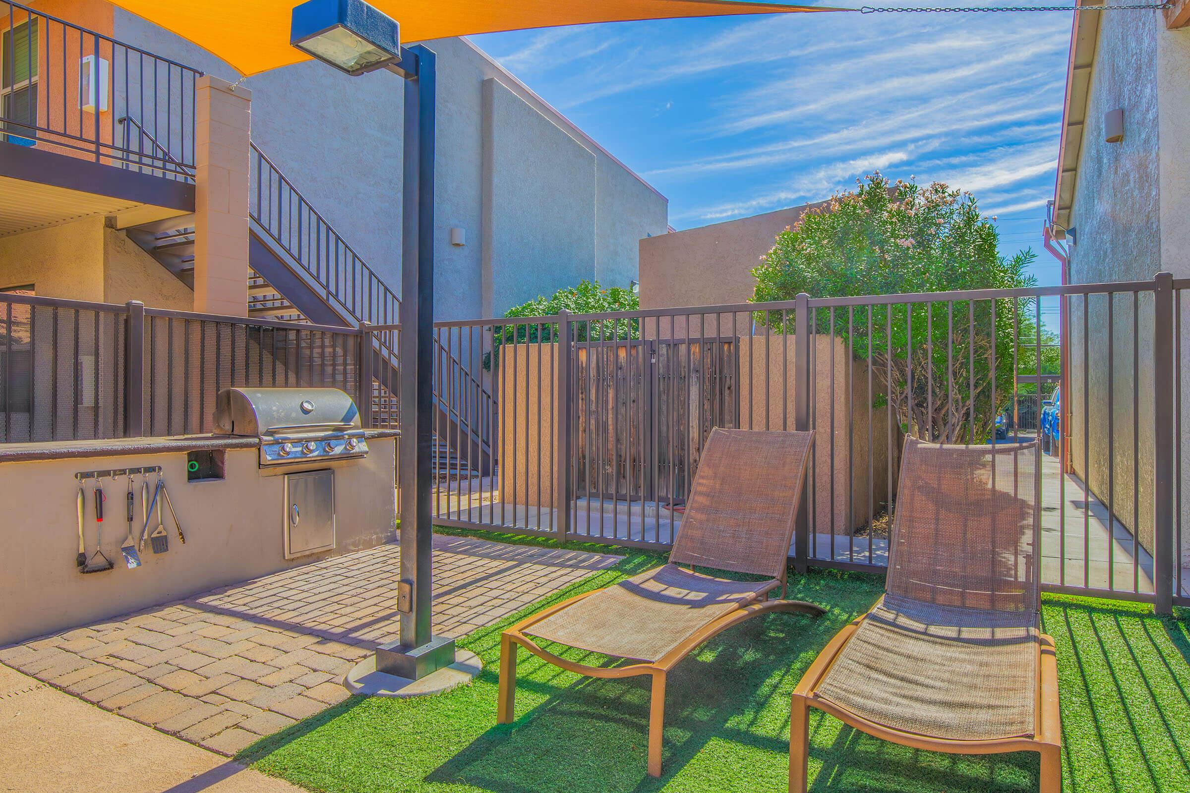 A cozy outdoor patio area featuring two lounge chairs and a grill, surrounded by a fence. The space is shaded by an umbrella, with a clear blue sky overhead. The flooring consists of pavers and artificial grass, creating a welcoming environment for relaxation and outdoor cooking.