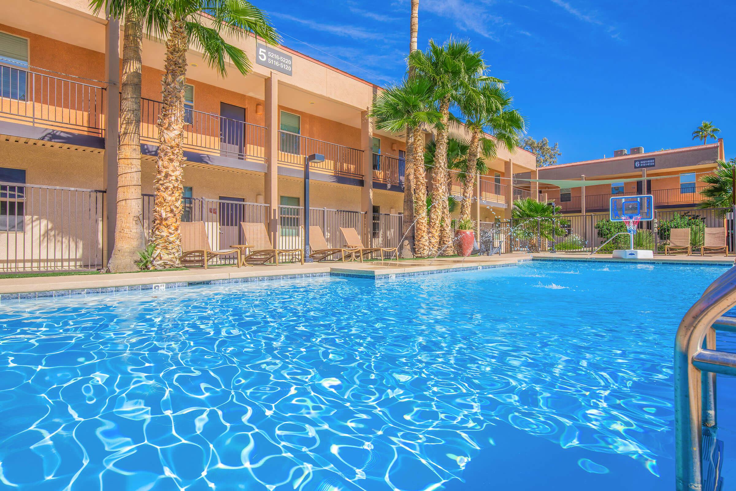 A crystal-clear swimming pool surrounded by palm trees and lounge chairs, with a basketball hoop visible in the background. The pool reflects the bright blue sky and the colorful building facade features balconies overlooking the area. Ideal for relaxation and recreation.