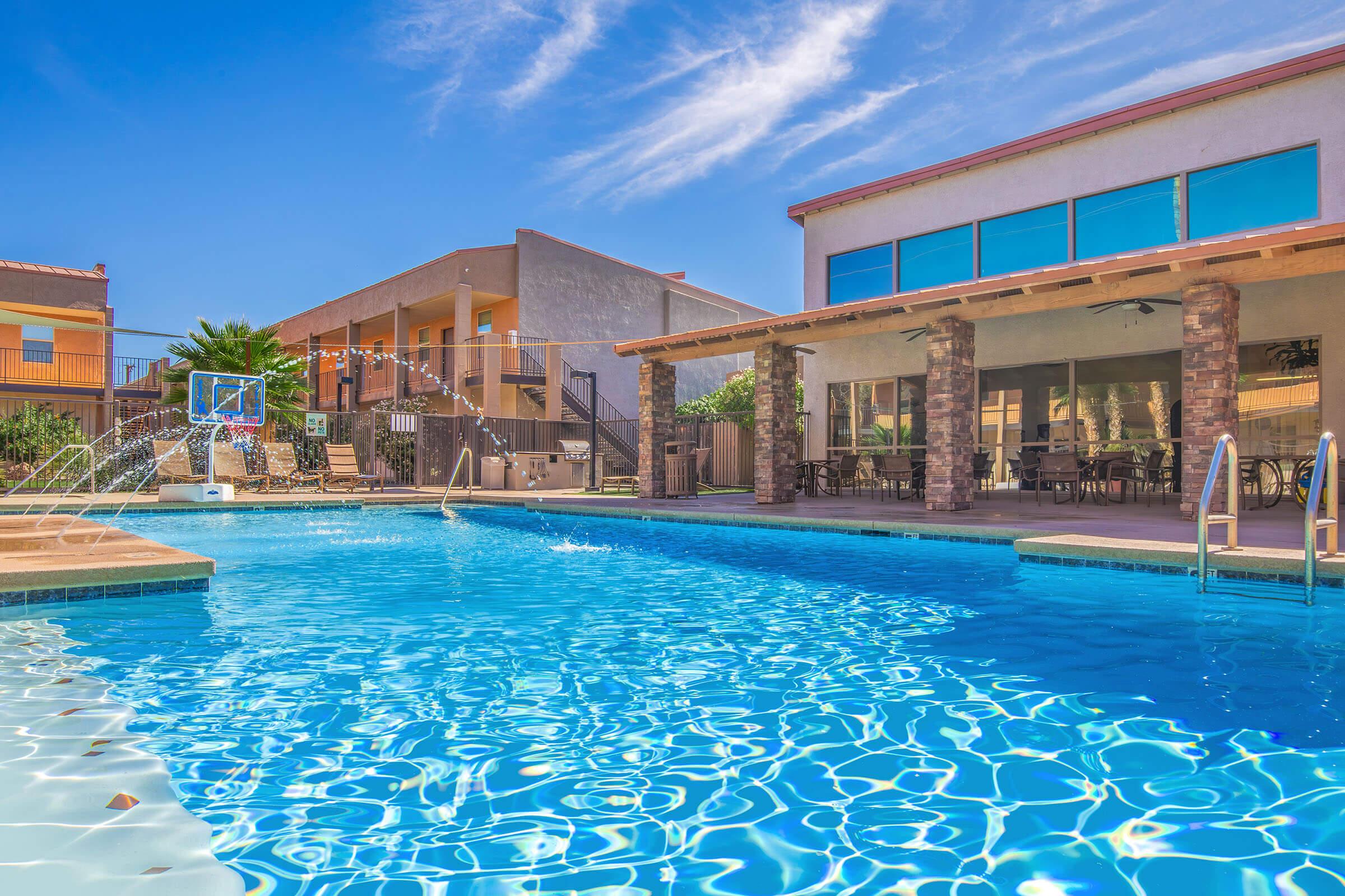 A bright, inviting swimming pool reflects the clear blue sky, surrounded by lush greenery. In the background, two buildings with outdoor seating areas and an elevated deck can be seen. The pool features a basketball hoop and a gentle water fountain, creating a relaxed and recreational atmosphere.