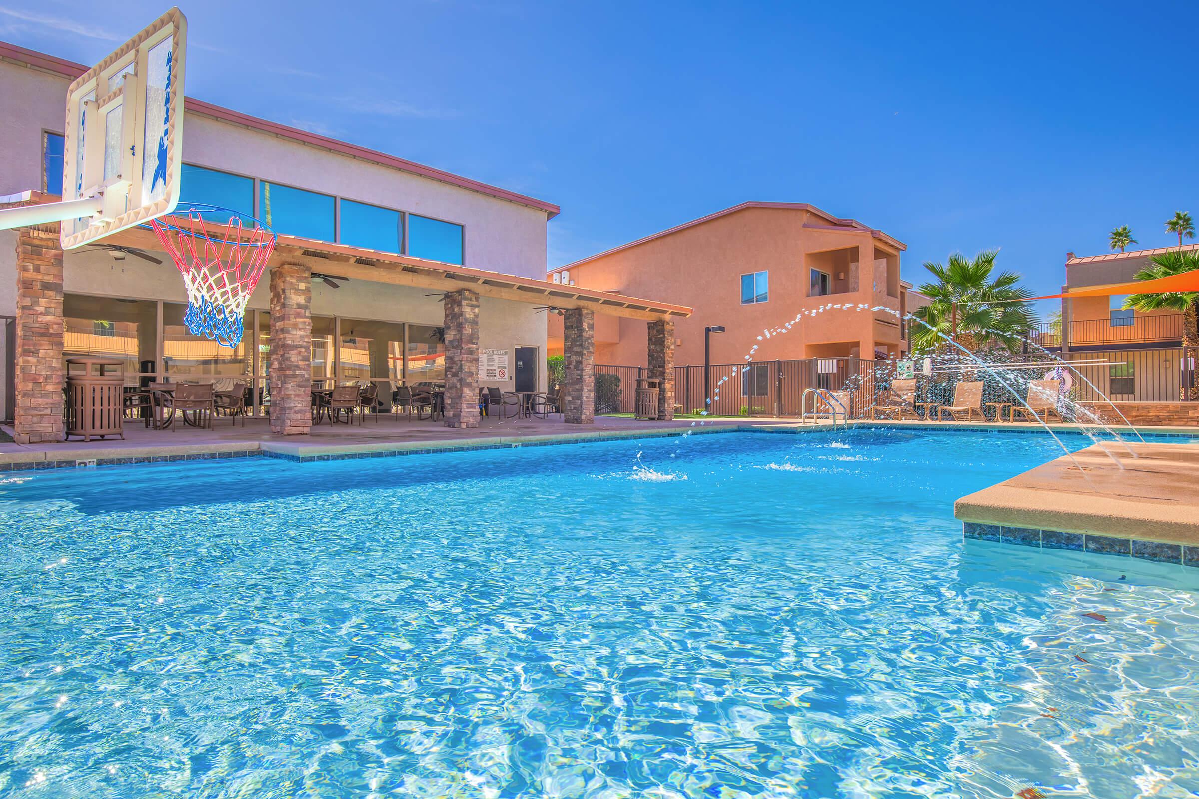 A sparkling blue swimming pool with water splashing from fountains. In the foreground, there is a basketball hoop beside the pool. Surrounding the pool are sunlit buildings with outdoor seating areas, palm trees, and clear blue skies, creating a vibrant and inviting atmosphere.