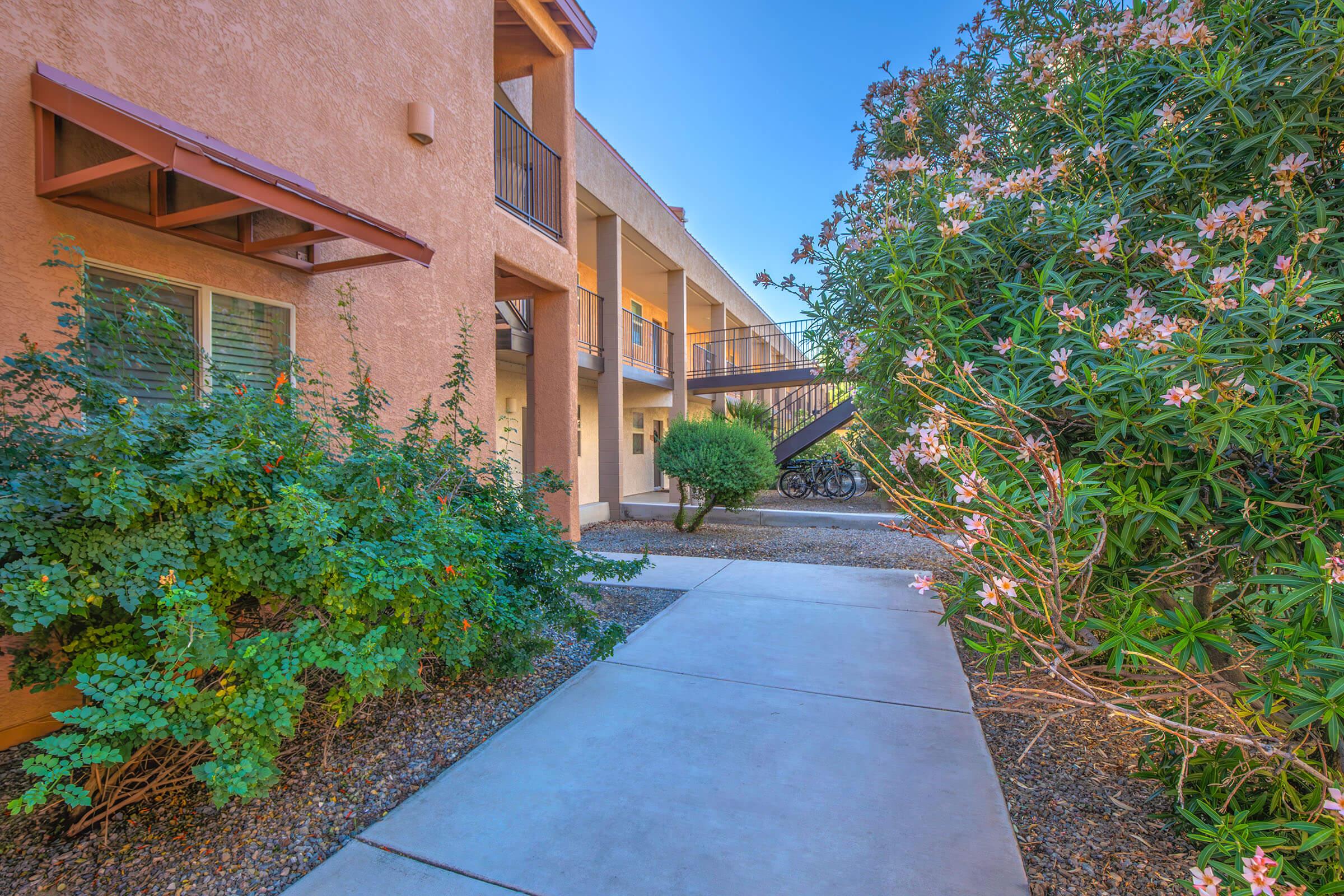 A pathway between two buildings, bordered by green shrubs and flowering plants. In the background, a staircase leads to the upper level, and bicycles are parked nearby. The scene is bathed in bright daylight, creating a welcoming atmosphere.