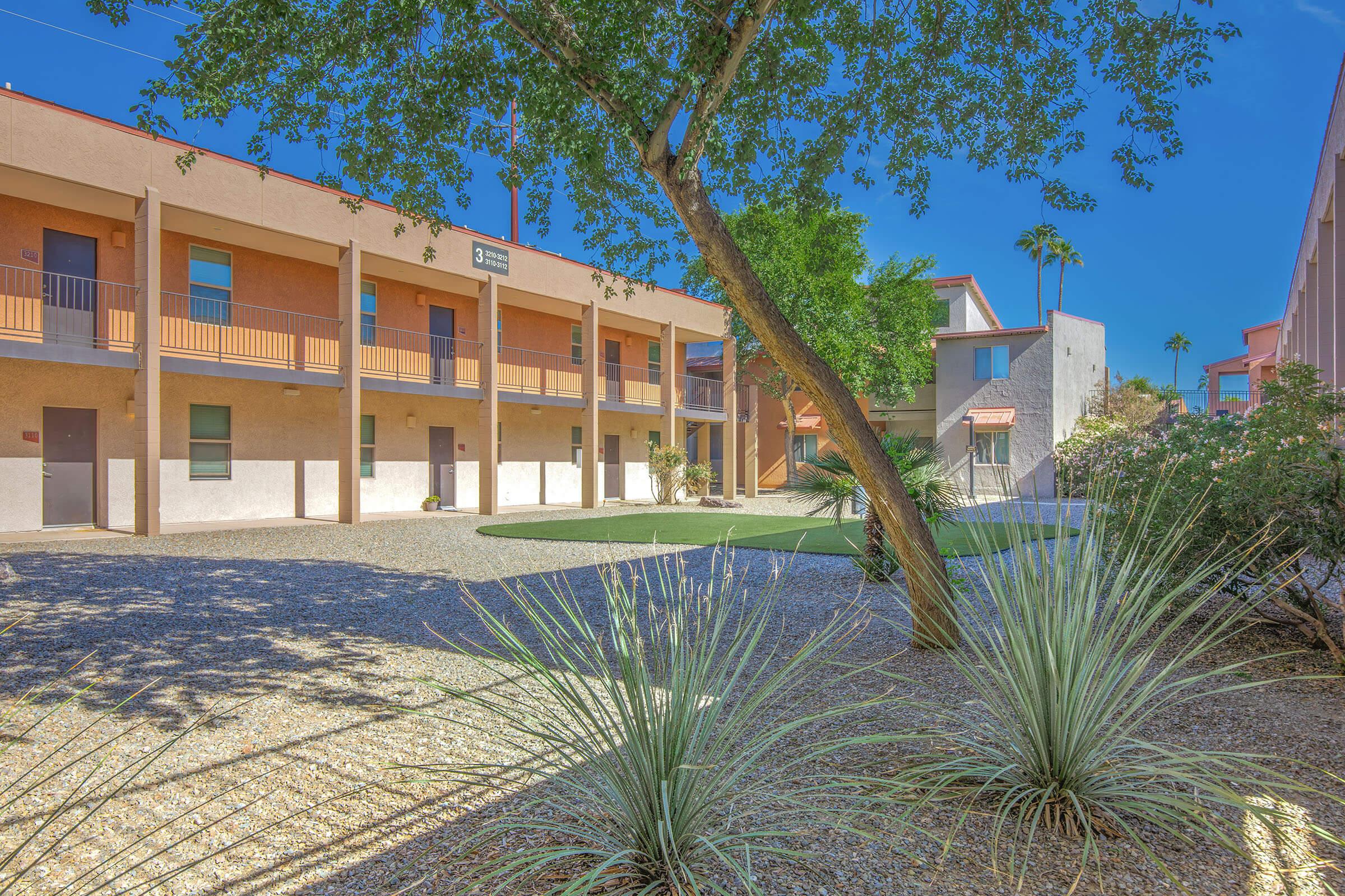 A serene courtyard scene featuring two-story buildings with balconies, surrounded by desert landscaping including gravel, plants, and a tree. The sky is clear and blue, creating a bright atmosphere.