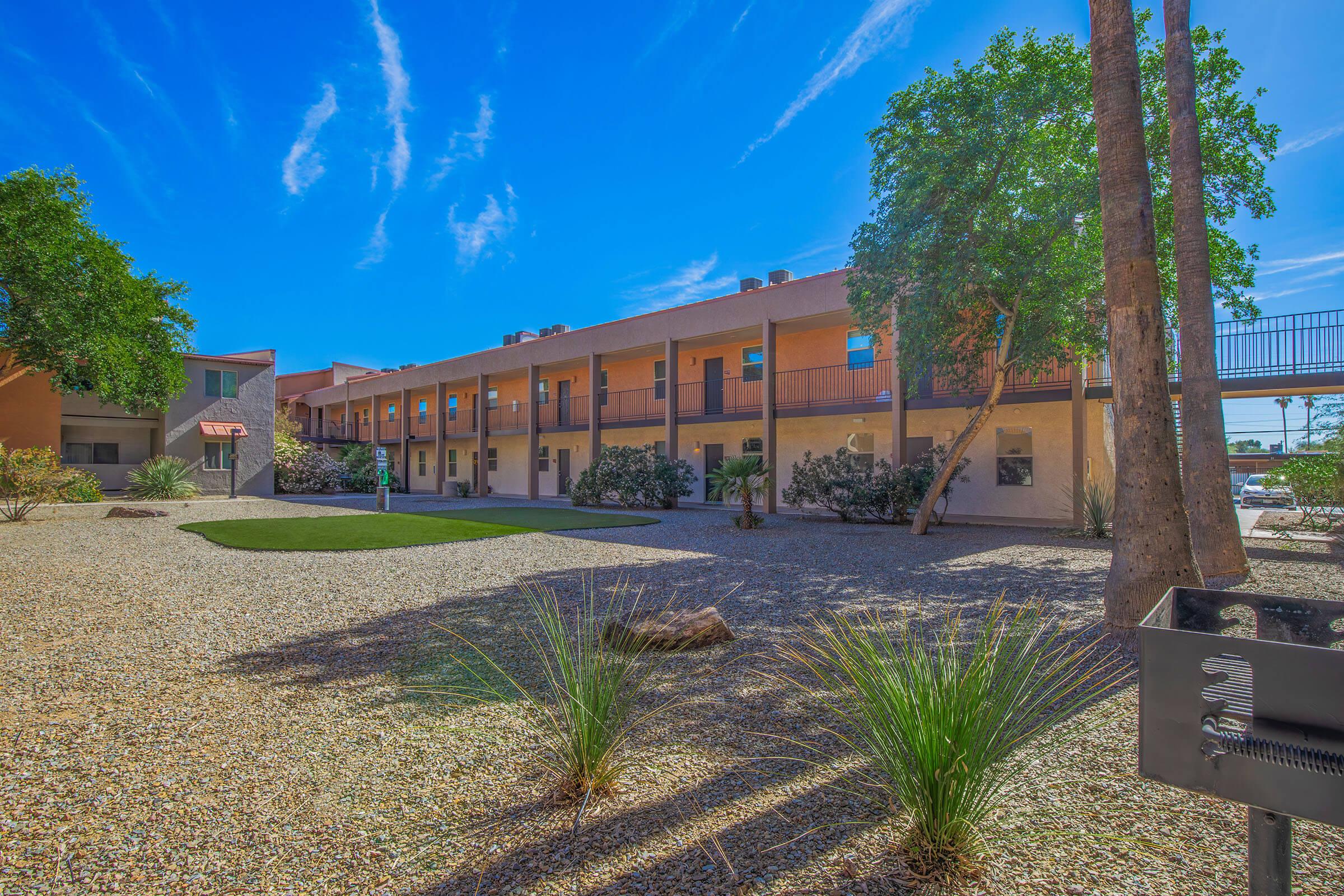 Outdoor view of a modern two-story building with a desert landscape. The foreground features gravel and desert plants, while the building has a light-colored facade with balconies. Bright blue sky with wispy clouds enhances the inviting atmosphere.