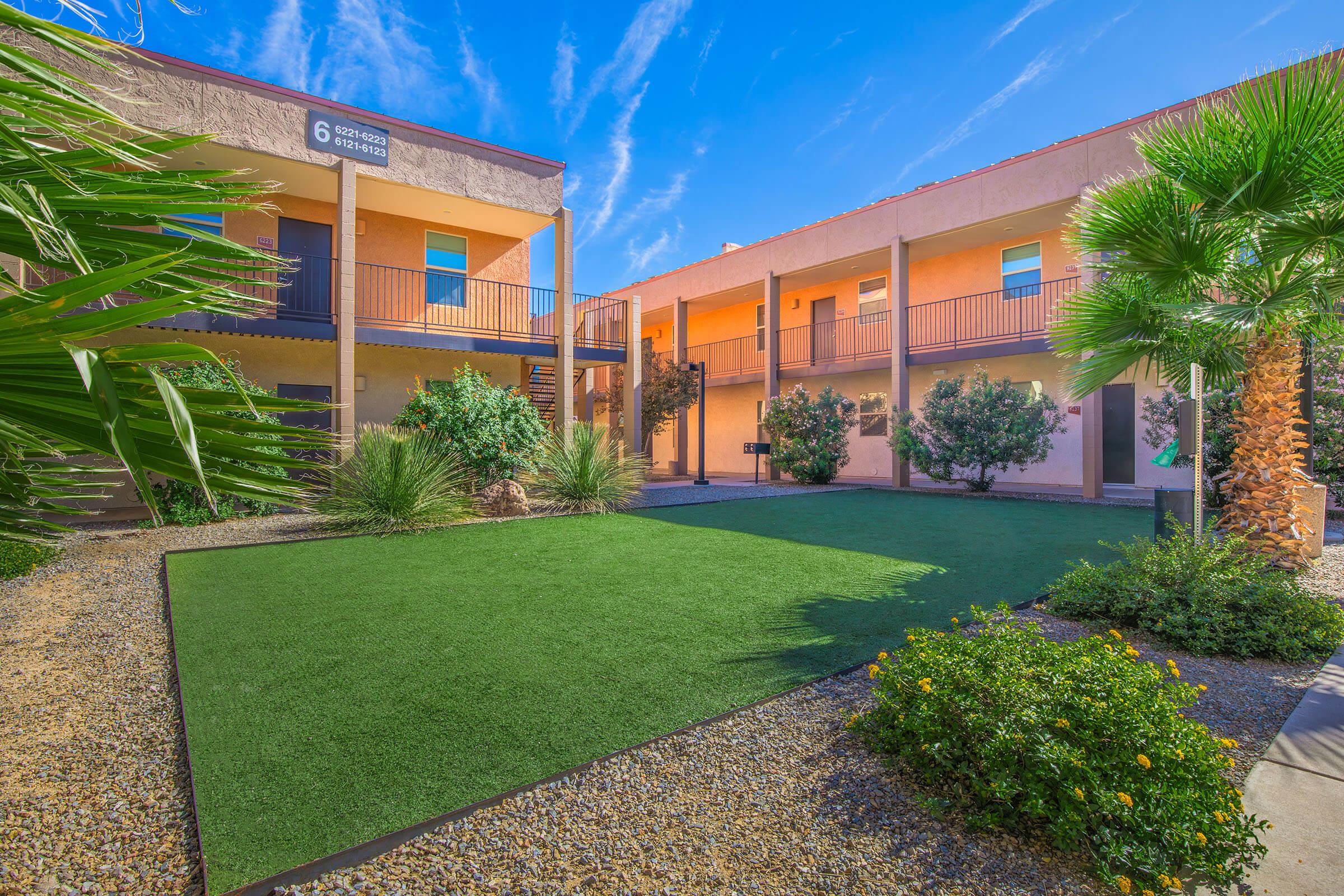 A view of the courtyard of a multi-level building featuring green grass-like landscaping, palm trees, and shrubs. The two-story structure has balconies and numbered doors, with a clear blue sky above. The area is designed for both aesthetic appeal and relaxation.