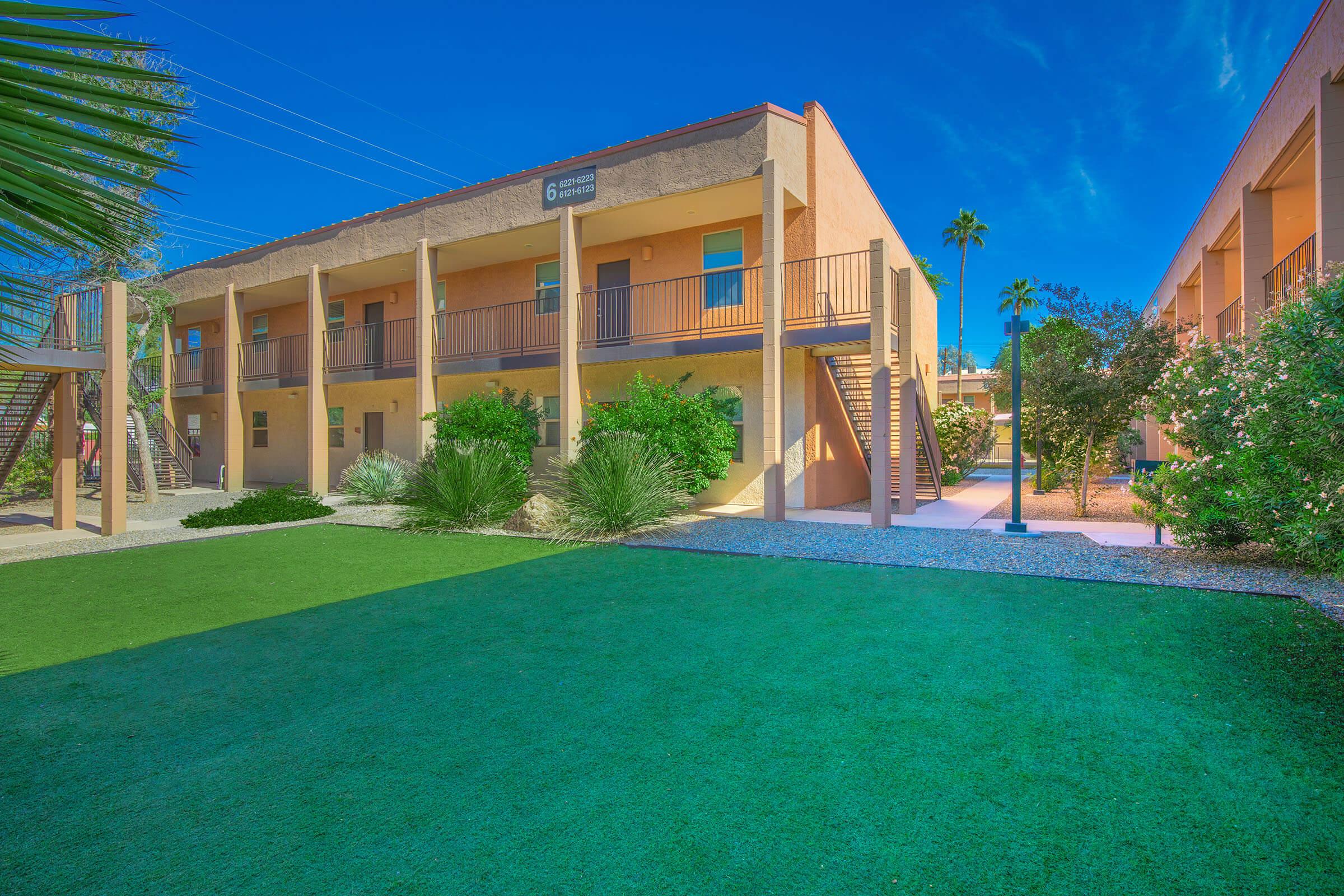 A sunny outdoor view of a two-story building with balconies. Lush greenery and palm trees surround the space, with a well-maintained lawn and pathways leading to the entrance. The clear blue sky adds to the vibrant atmosphere of the setting.