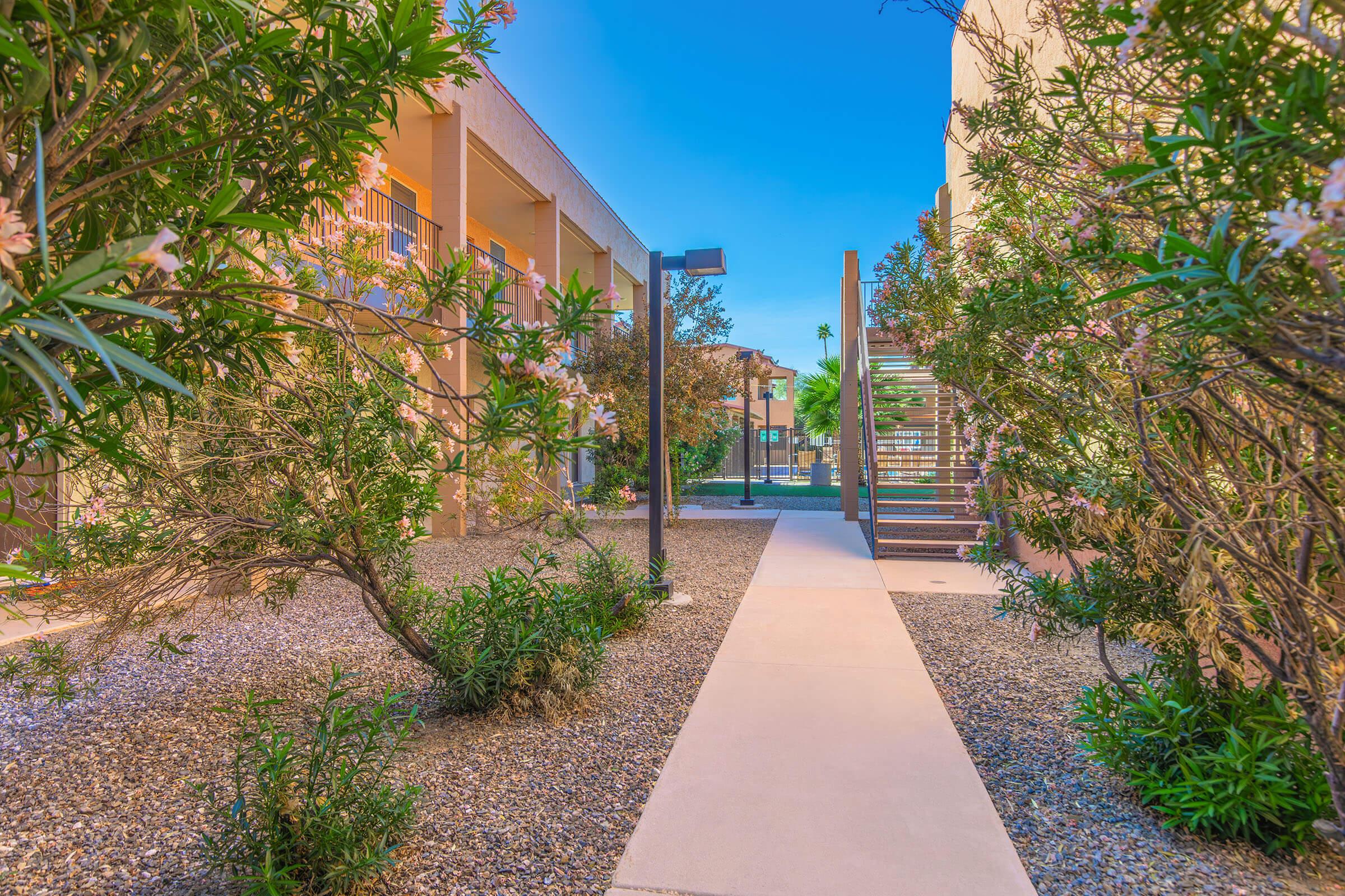 A pathway surrounded by lush greenery and flowering shrubs leads through a landscaped area, flanked by modern buildings. A metal staircase ascends to a second level in the background, with clear blue skies above, creating a serene outdoor environment.
