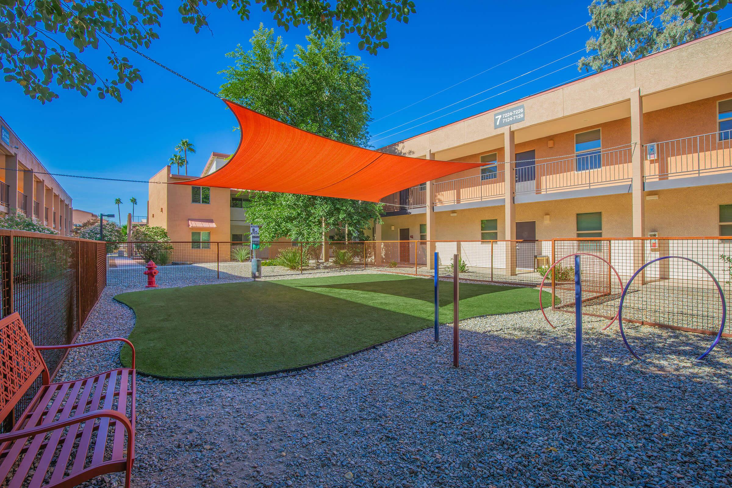 Vibrant outdoor space featuring a green lawn area shaded by an orange canopy, surrounded by a gravel path. In the background, two stories of a building with balconies can be seen, along with a fire hydrant and playful structures like hoops. Bright blue sky and greenery complete the scene.