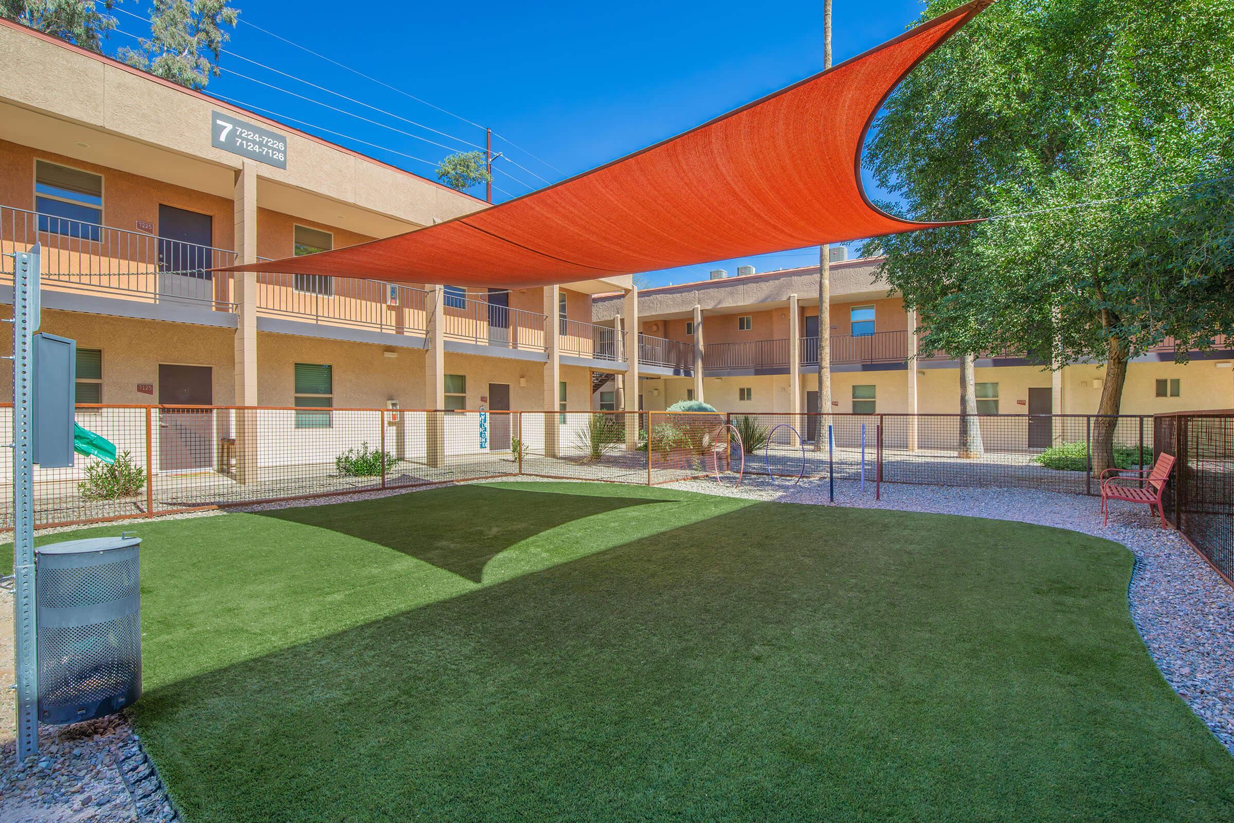 A well-maintained outdoor area with synthetic grass, shaded by a large red sail. Surrounding the space are two-story buildings with balconies. There are playground equipment and trees, creating a pleasant environment for relaxation and play. The area is enclosed by a fence, ensuring safety for children and pets.