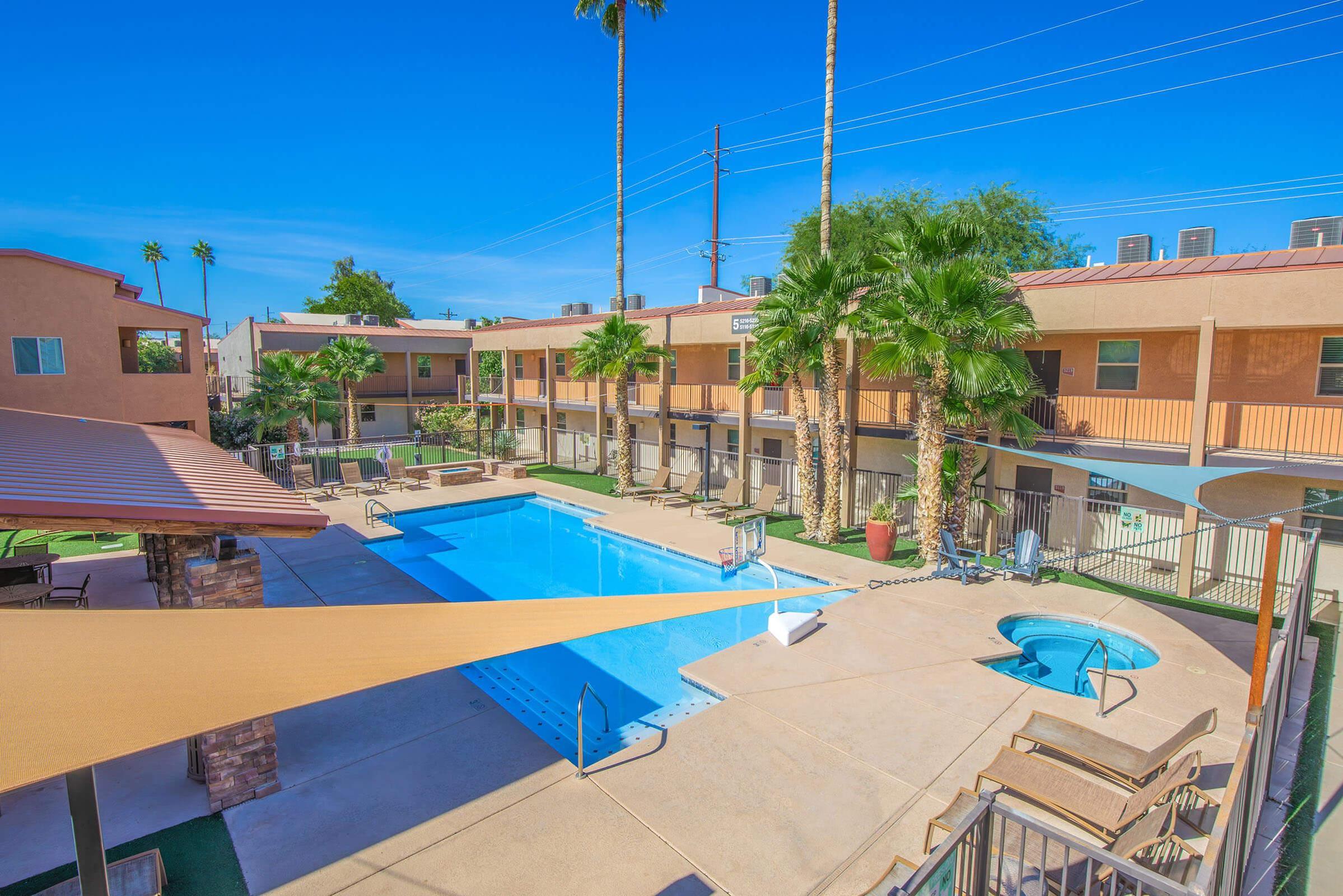A scenic view of a swimming pool area surrounded by palm trees, with lounge chairs and a hot tub. The pool area is flanked by two-story buildings, featuring a warm color palette. A clear blue sky and power lines are visible in the background, creating a bright and inviting atmosphere.