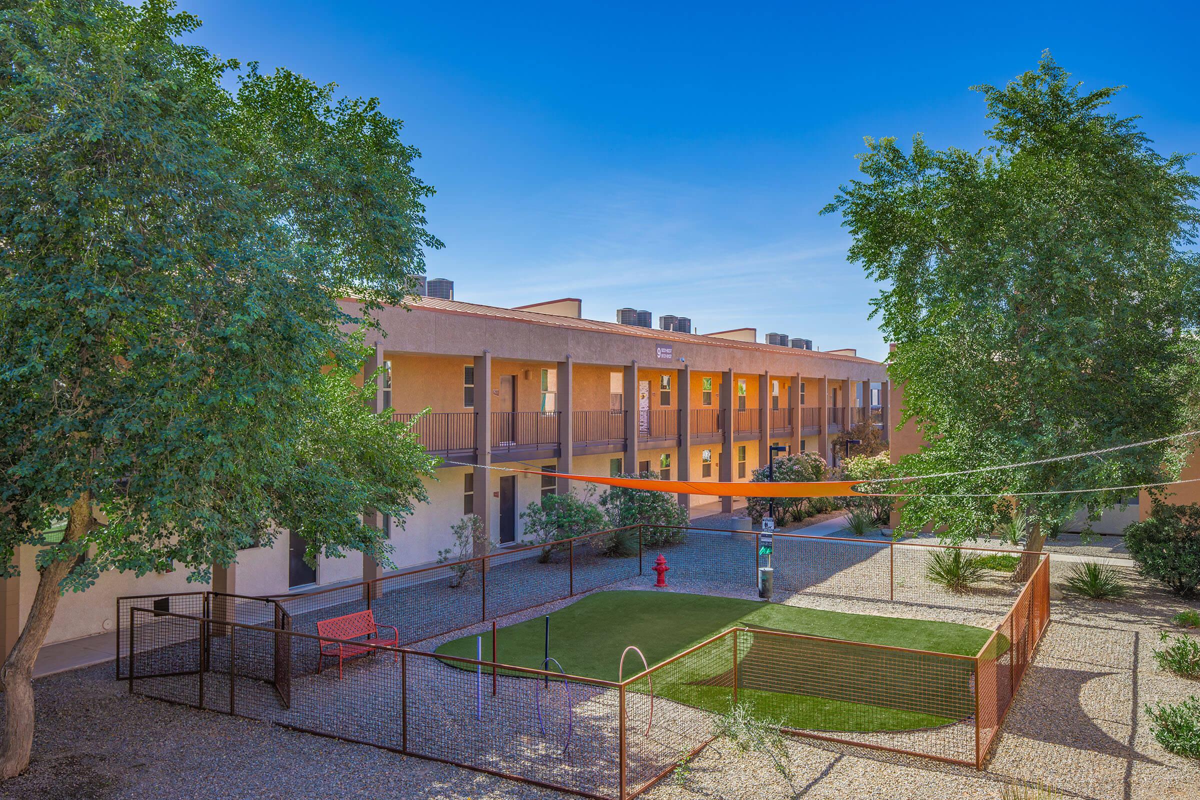 A sunny courtyard scene featuring apartment buildings with a bright blue sky. In the foreground, there is a green lawn enclosed by a low fence, with a bench and a fire hydrant. The area is surrounded by trees and landscaped with gravel and plants, creating a peaceful outdoor environment.