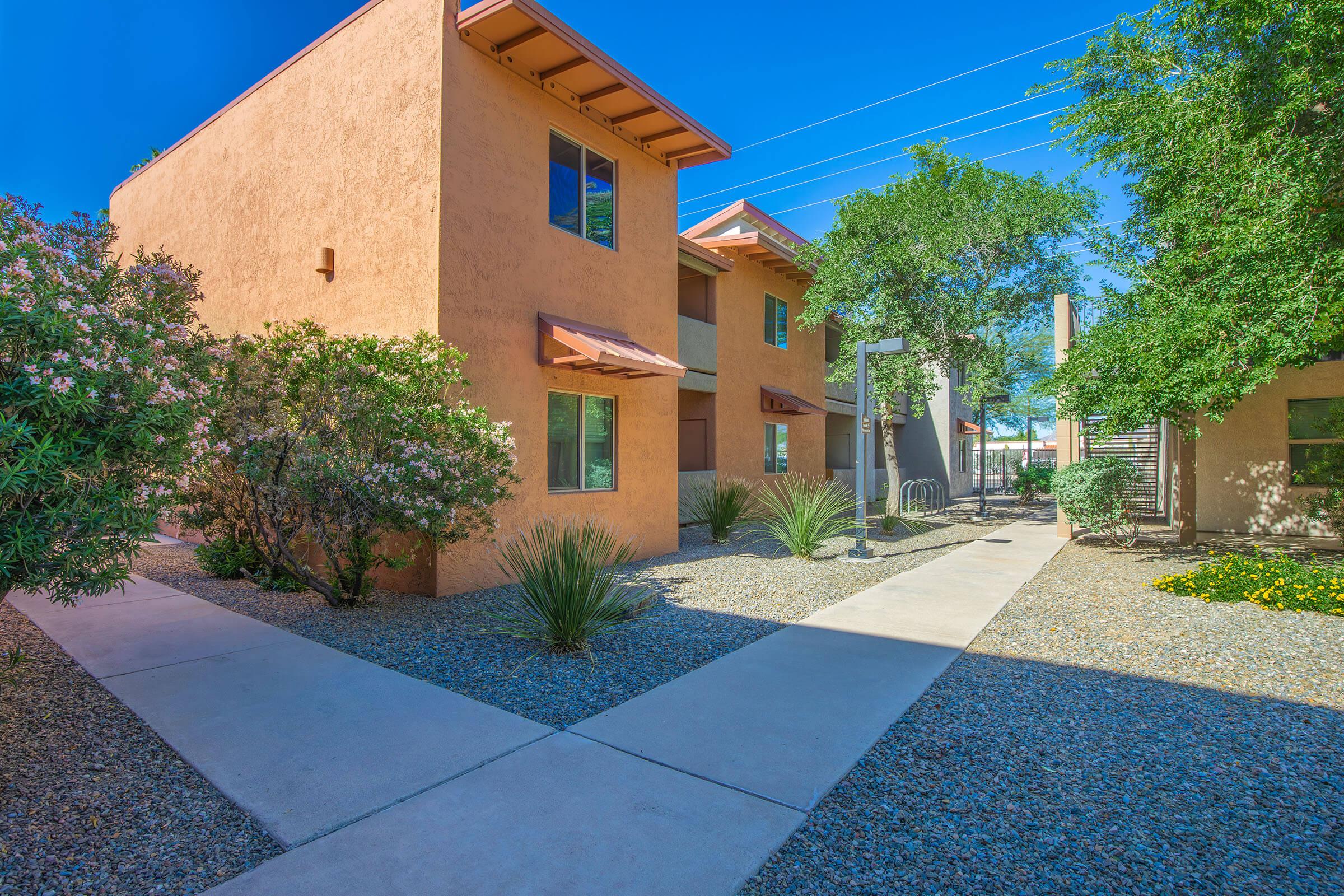 A landscaped courtyard area with two brown stucco buildings on either side. The path leads through gravel and surrounded by desert plants and small trees. Bright blue sky overhead, providing a sunny and inviting atmosphere.