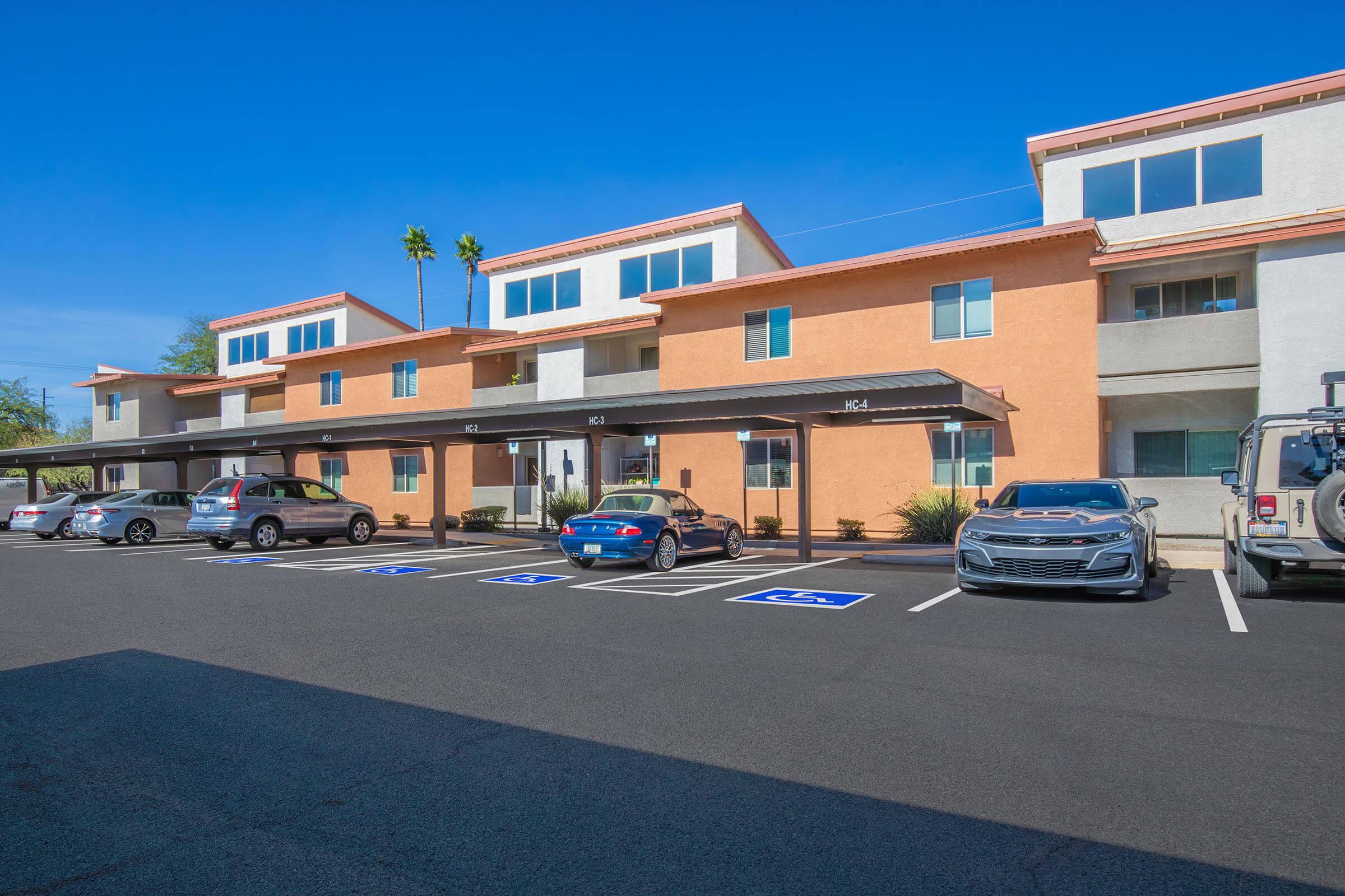 A row of modern apartment buildings with a mix of orange and gray exteriors. Several cars are parked in front, including a blue sedan and a silver sports car. The sky is clear and blue, with a few palm trees in the background. Accessible parking spaces are visible in the foreground.