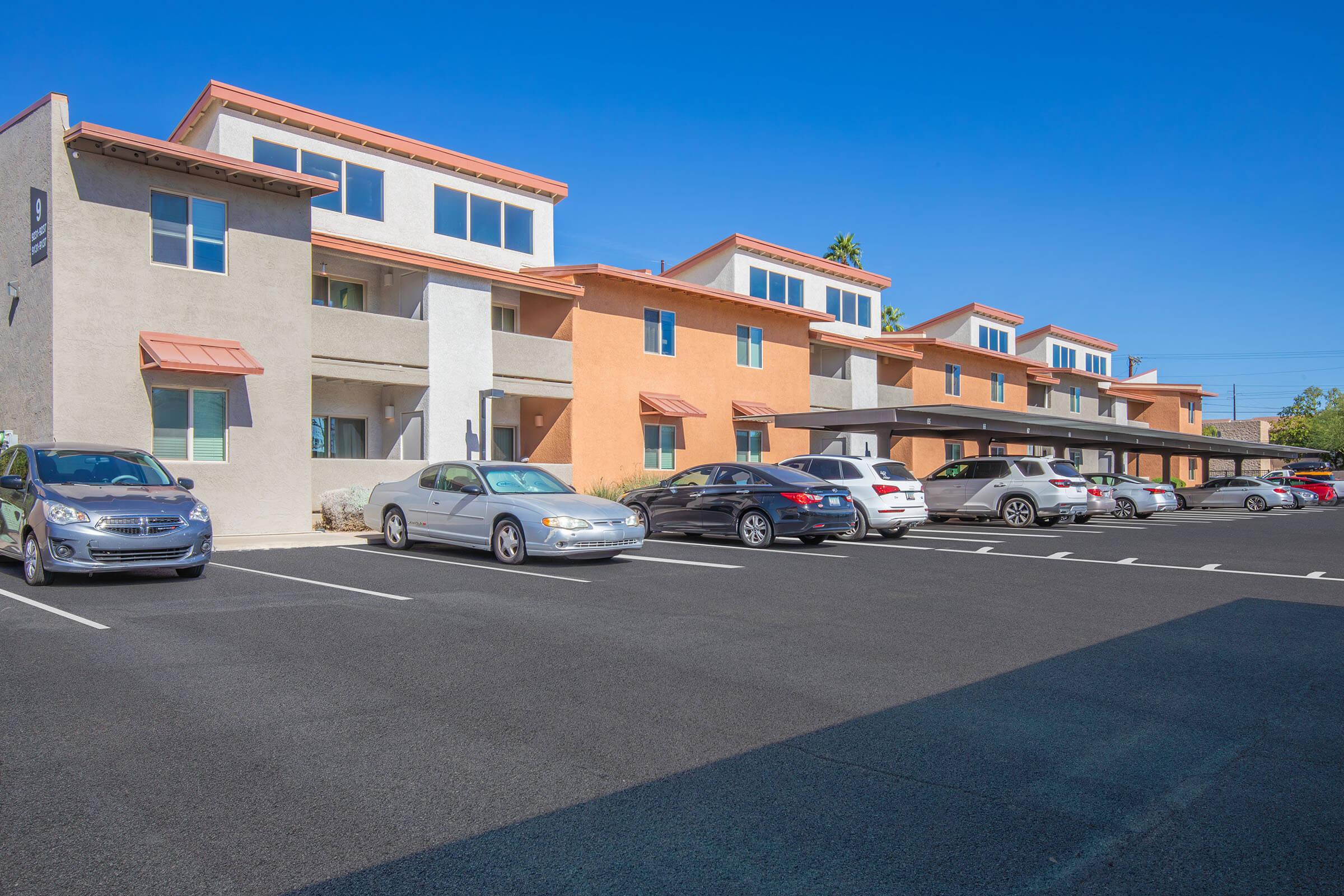 A row of residential buildings featuring modern architecture, with multiple parked cars in a well-maintained parking lot. The buildings have different colors and styles, under a clear blue sky.