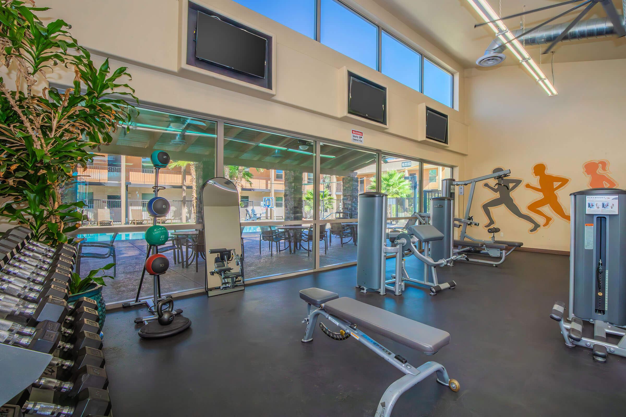 A modern gym interior featuring workout equipment like benches and weight machines. Large windows allow natural light to illuminate the space, and there are TVs mounted on the walls. In the background, a pool area can be seen through glass doors, and there are fitness-themed silhouettes on the wall.
