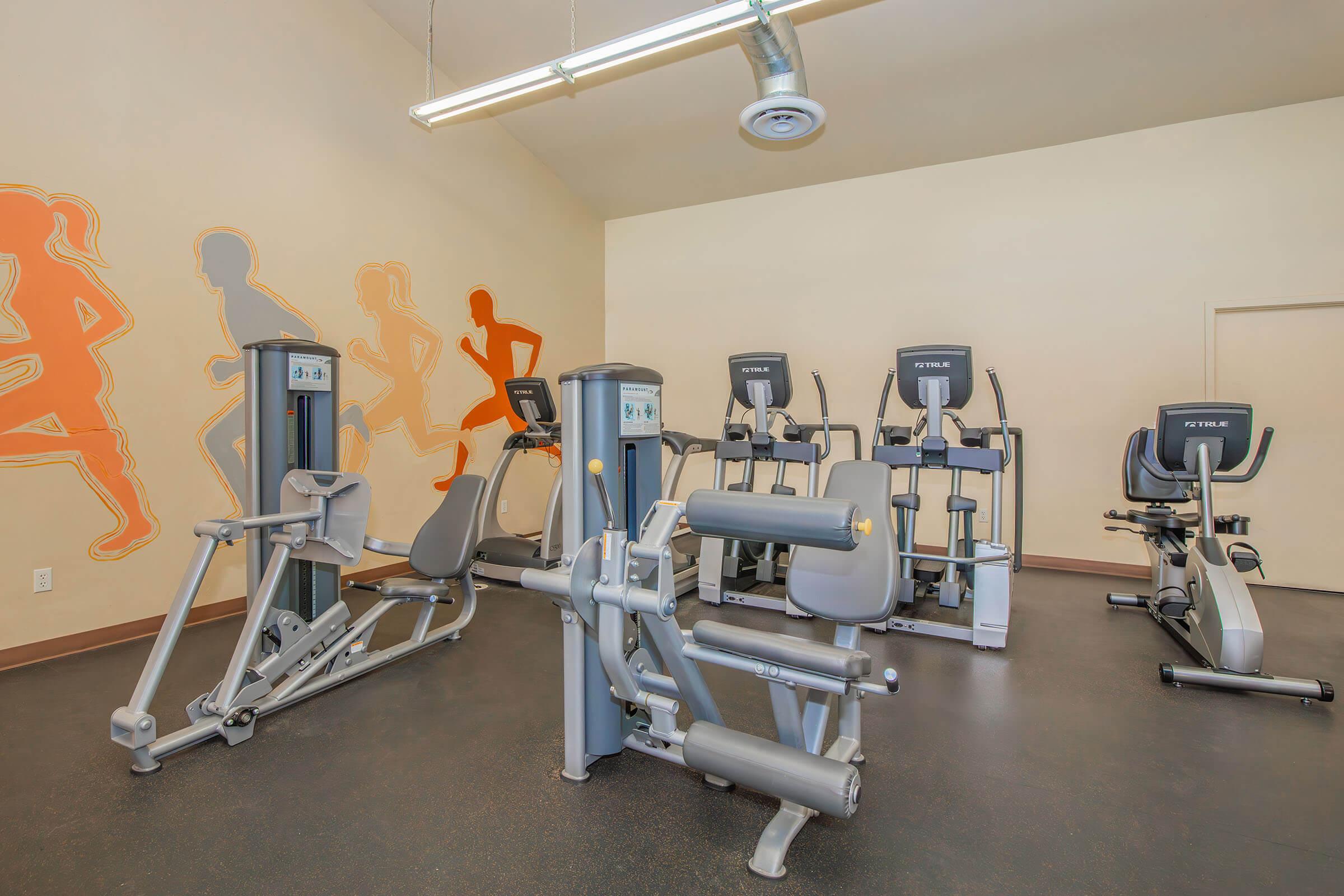 A fitness room featuring various exercise equipment, including stationary bikes and strength training machines. The walls are decorated with orange silhouettes of runners, creating an energetic atmosphere. The floor is dark, and there's overhead lighting for a bright environment.