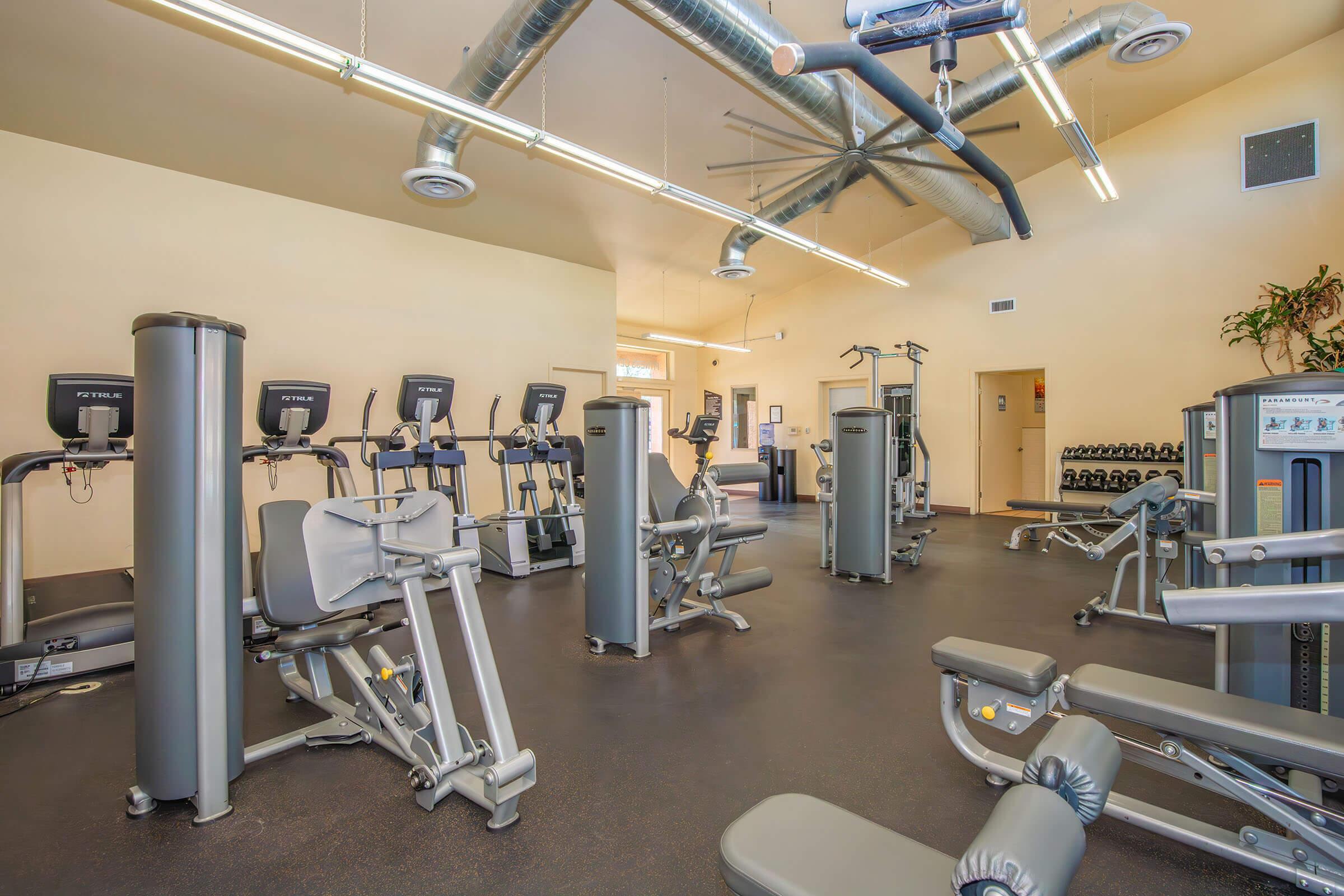 A spacious and well-lit gym interior featuring various exercise machines, including treadmills, weight machines, and benches. The room has a modern design with high ceilings, exposed ductwork, and natural light from windows. In the background, there are free weights neatly arranged on a rack.