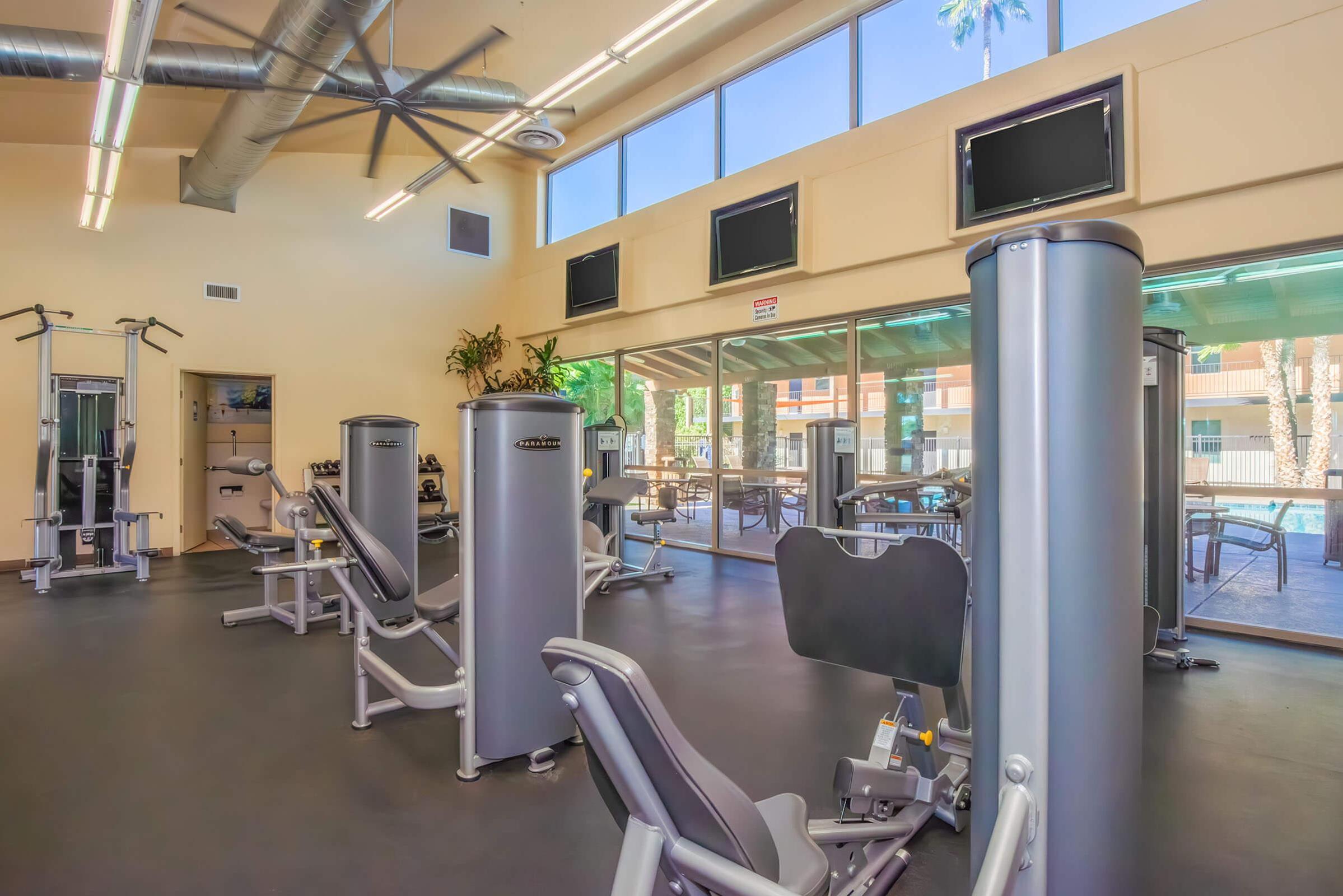 A bright, modern gym with various exercise machines and weights. Large windows allow natural light, offering a view of an outdoor pool area. The space is well-maintained, featuring a clean floor and a ceiling fan for ventilation. Fitness equipment is strategically placed for optimal workout flow.