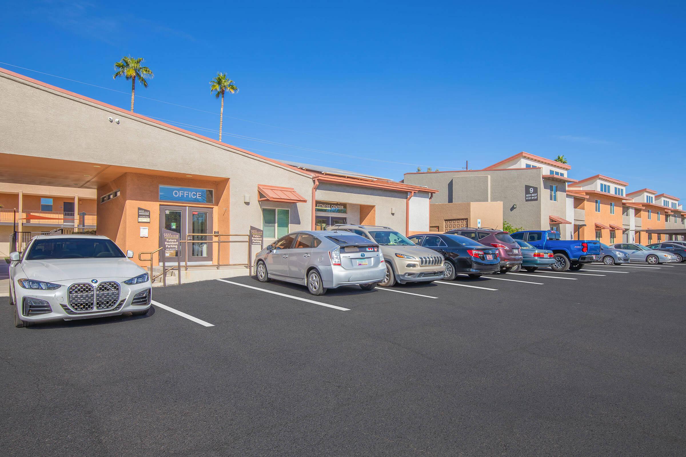 A parking lot with several cars parked in front of a building labeled "OFFICE." The building features a light-colored exterior, an awning above the entrance, and palm trees in the background against a clear blue sky. Additional buildings can be seen in the distance.