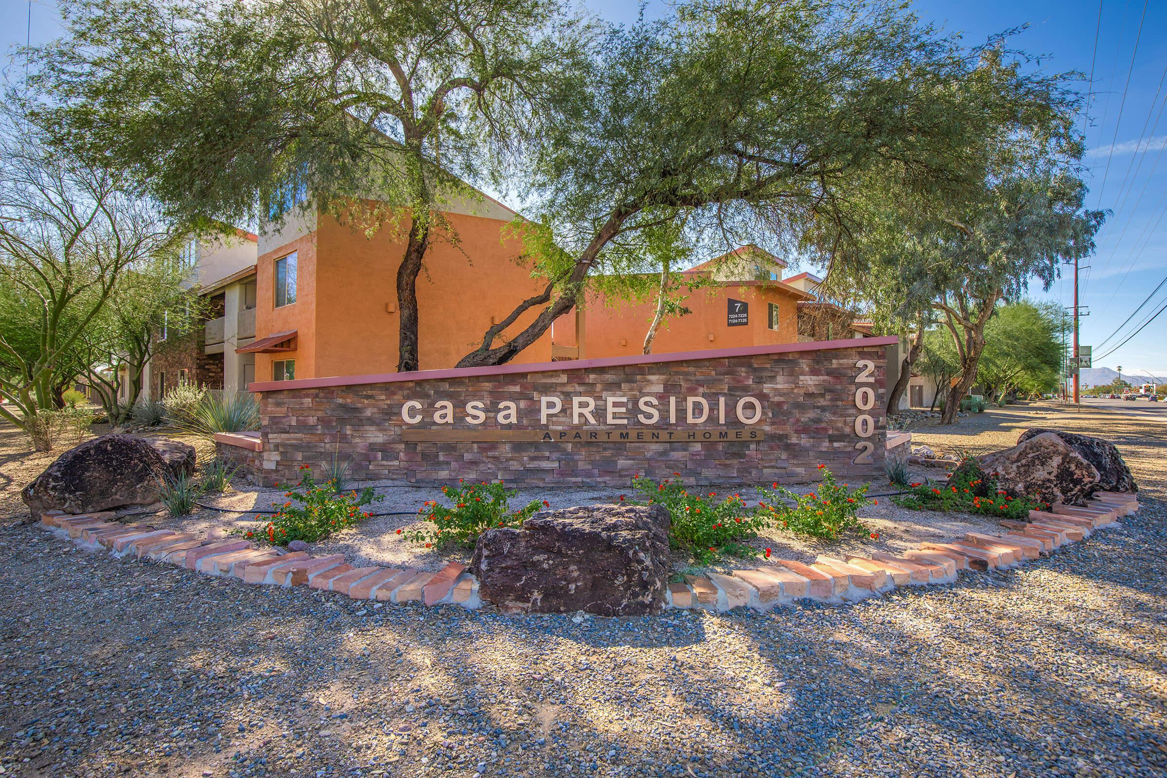 Sign for Casa Presidio Apartment Homes, featuring modern lettering on a stone structure surrounded by desert landscaping, including rocks and plants. The background shows a colorful building under a clear blue sky. The location is likely in a sunny, arid environment.