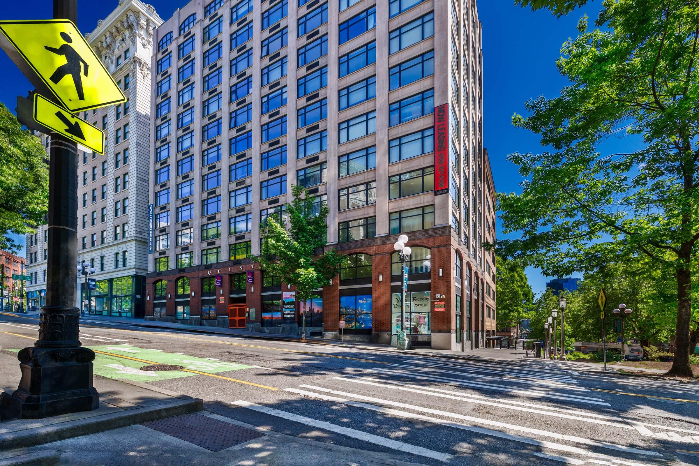 A city street view showcasing a multi-story building with large windows, surrounded by trees and pedestrian traffic signals. Clear blue sky and bright sunlight enhance the scene, while the street is marked with bike lanes and crosswalks, indicating a busy urban environment.