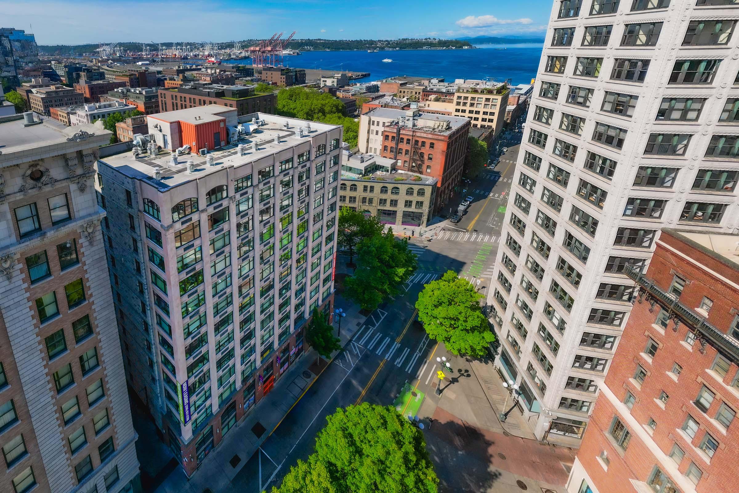 Aerial view of a cityscape featuring tall buildings, tree-lined streets, and a glimpse of water in the background. The scene captures a mix of modern and historic architecture, with a vibrant urban environment under a clear blue sky.