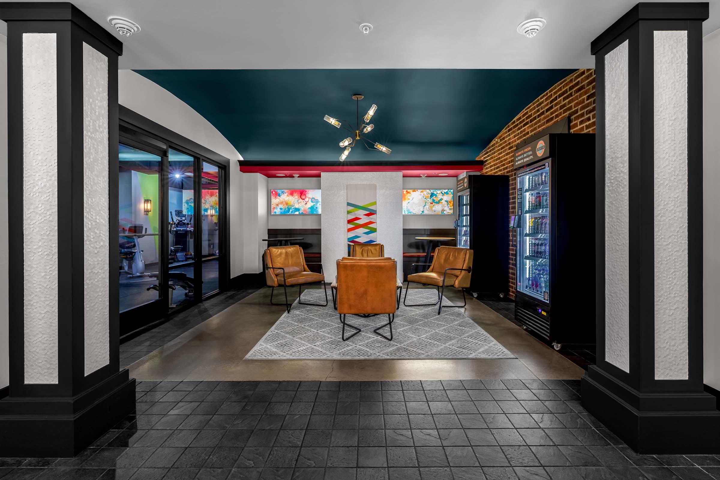 A modern lounge area featuring two brown leather chairs and a geometric rug. In the background, colorful artwork hangs on the walls. A snack and drink vending machine is visible on the right, while glass doors lead to another space. The ceiling is painted blue, and light fixtures add a stylish touch.