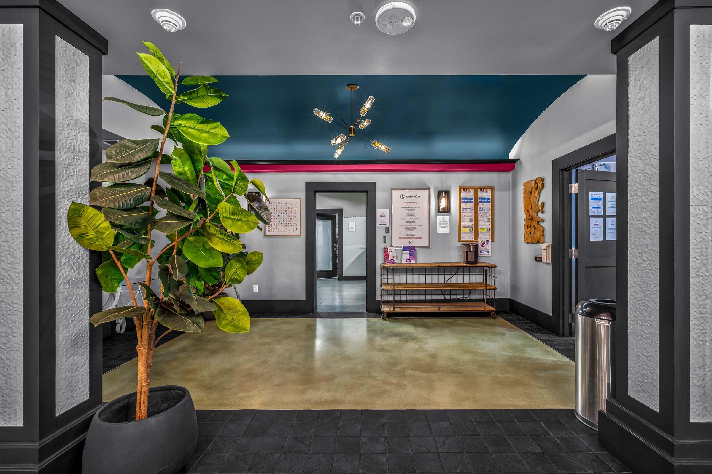 A spacious foyer with a large potted plant on the left, a modern light fixture hanging from the ceiling, and a variety of informational posters on the walls. There is a shelf with items for guests and a doorway leading to another area, all featuring a contemporary design and color scheme.