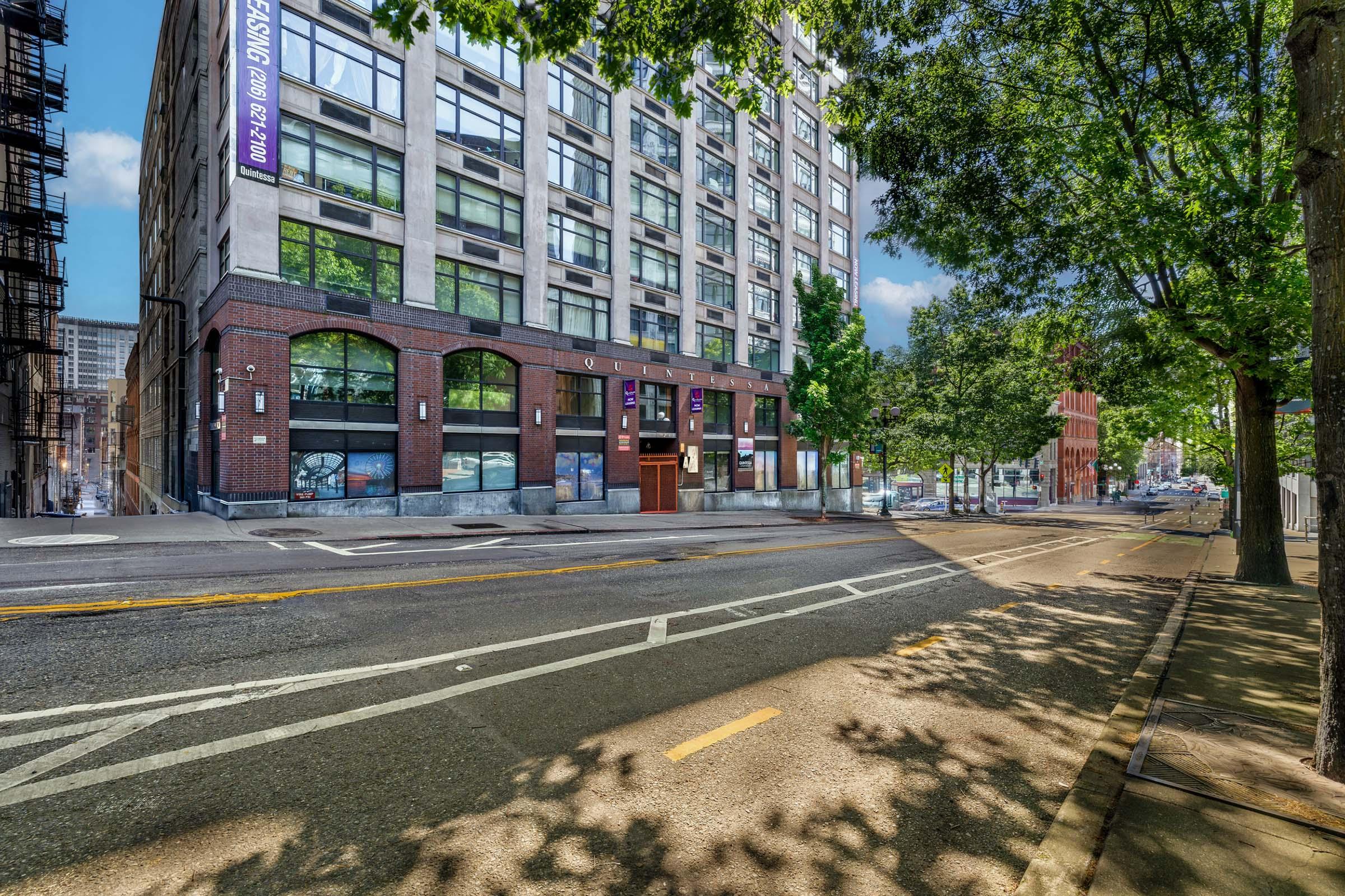 A modern multi-story building with large windows and a brick exterior is shown on a city street. Lush green trees line the sidewalk, and a few parked cars are visible. The scene is bright and sunny, giving a vibrant urban atmosphere.