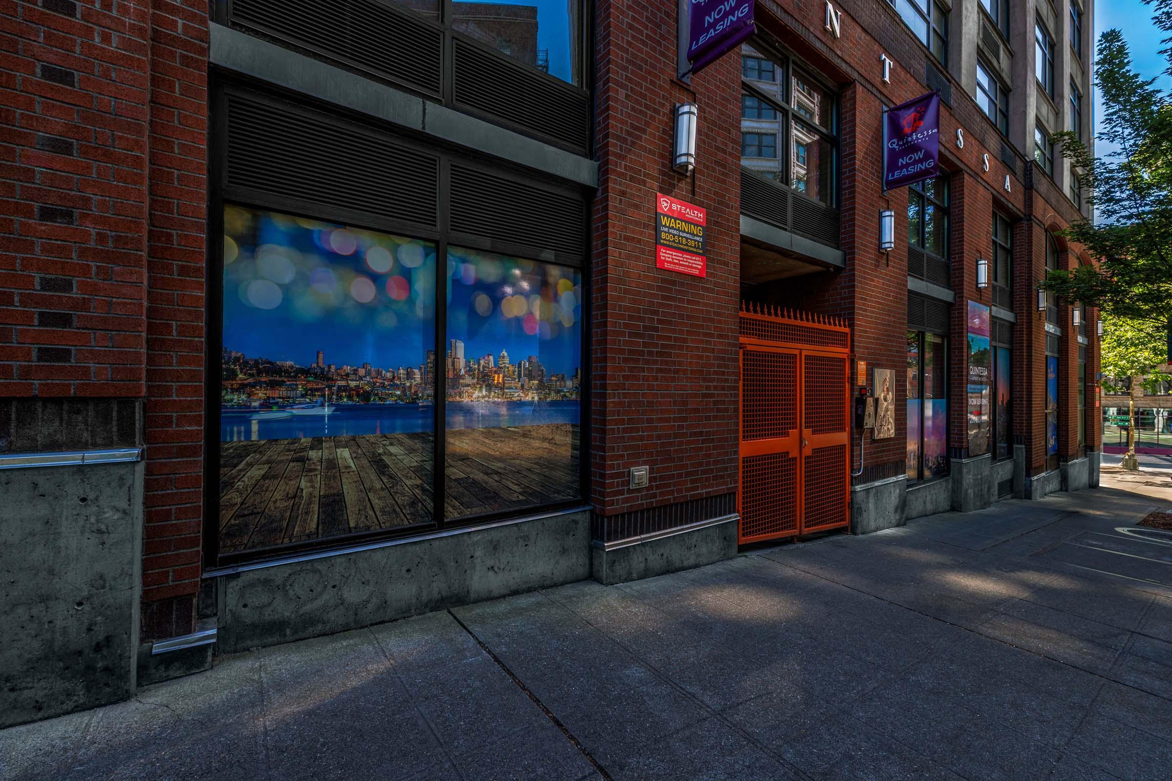 A cityscape view of a waterfront at night is displayed on a window, with colorful bokeh lights in the background. The setting features a brick building with metal accents, showcasing a modern urban environment. The sidewalk leads to the entrance, creating a welcoming atmosphere.
