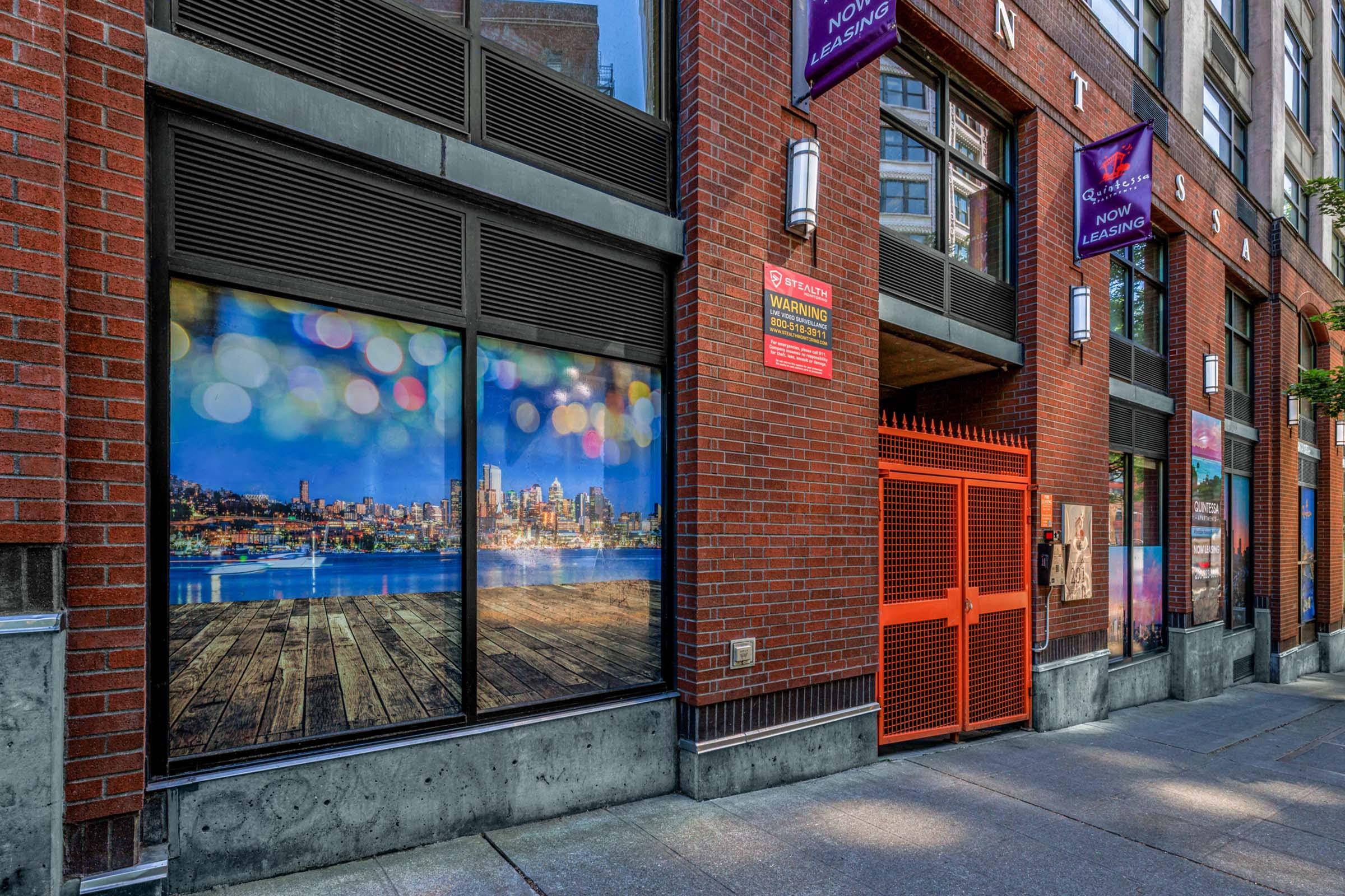 Exterior view of a brick building with large windows displaying a scenic city skyline and bokeh lights. The entrance features an orange gate, and various signs are posted on the wall. The setting suggests a modern urban environment.