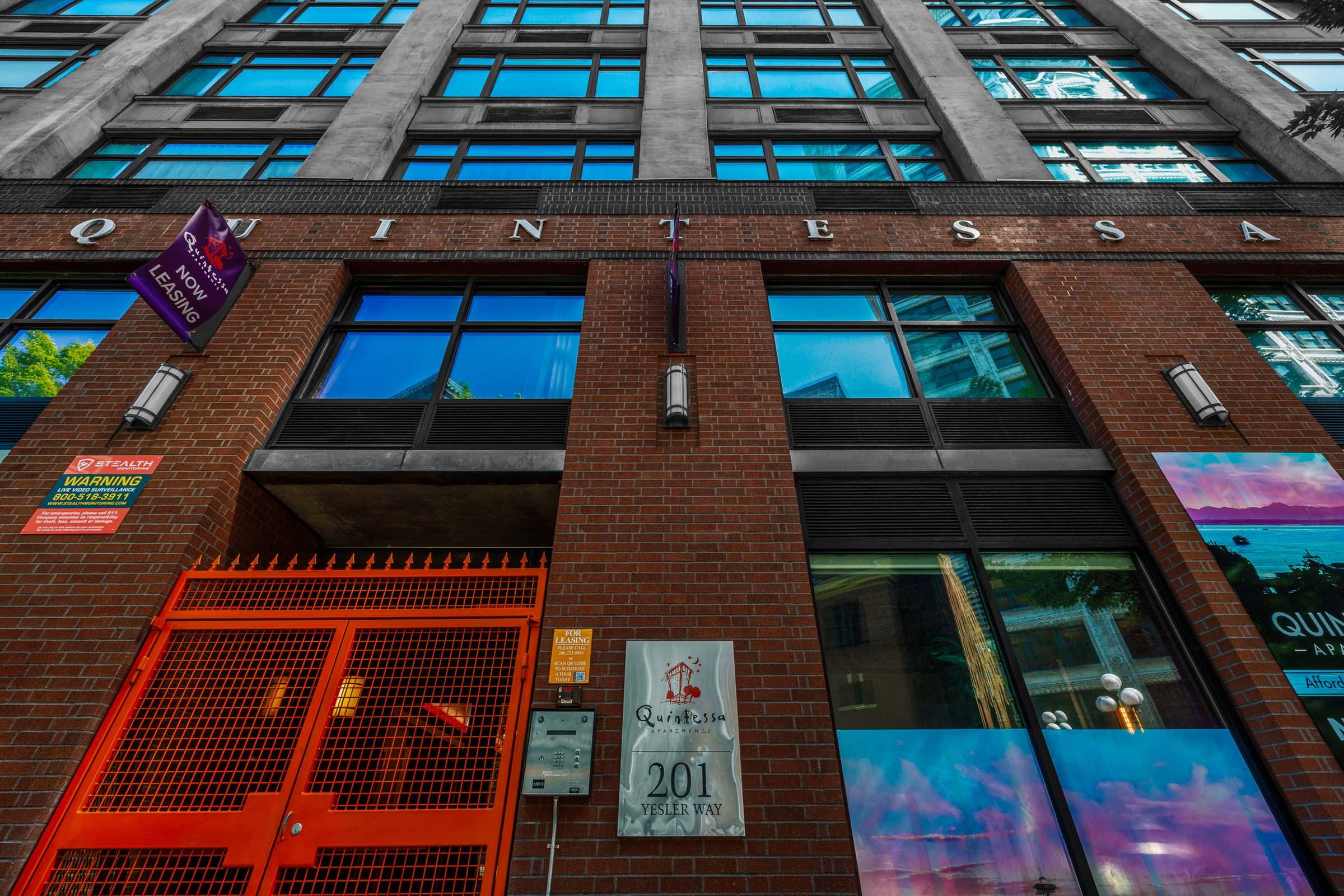 Image of a modern building facade featuring large windows and a red brick ground level. The entrance is highlighted with a red gate and a sign indicating the address "201". A banner hangs from the building with the word "Quintessa" visible above the entrance. The surroundings include vibrant colors and natural light.