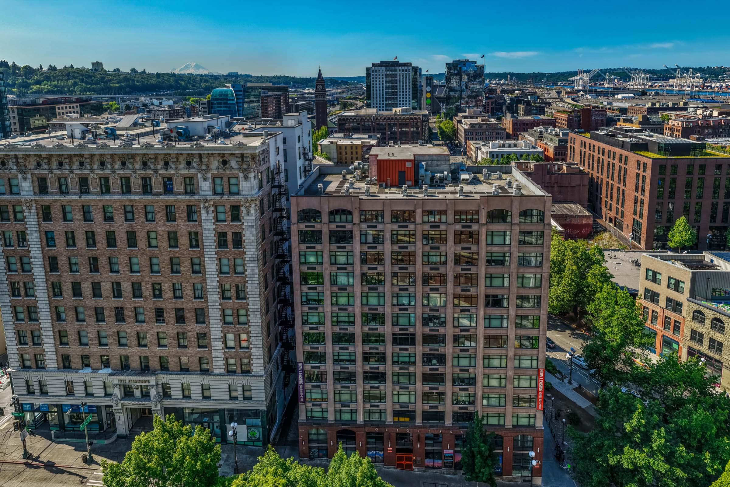 Aerial view of a cityscape featuring a mix of historic and modern buildings, tree-lined streets, and urban infrastructure under a clear blue sky. The scene includes a prominent tall structure in the background, indicating a vibrant downtown area with mixed architecture.