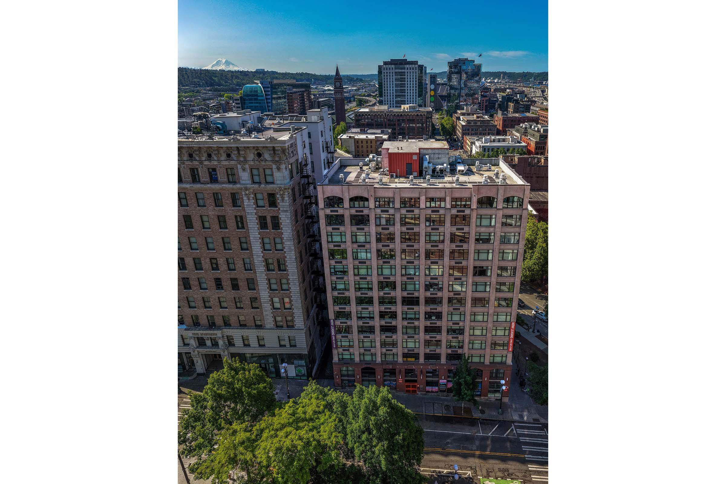 Aerial view of a cityscape featuring a mix of modern and historic buildings, with green trees in the foreground. The scene includes a clear blue sky and distant mountains, showcasing urban life and architecture in a vibrant setting.