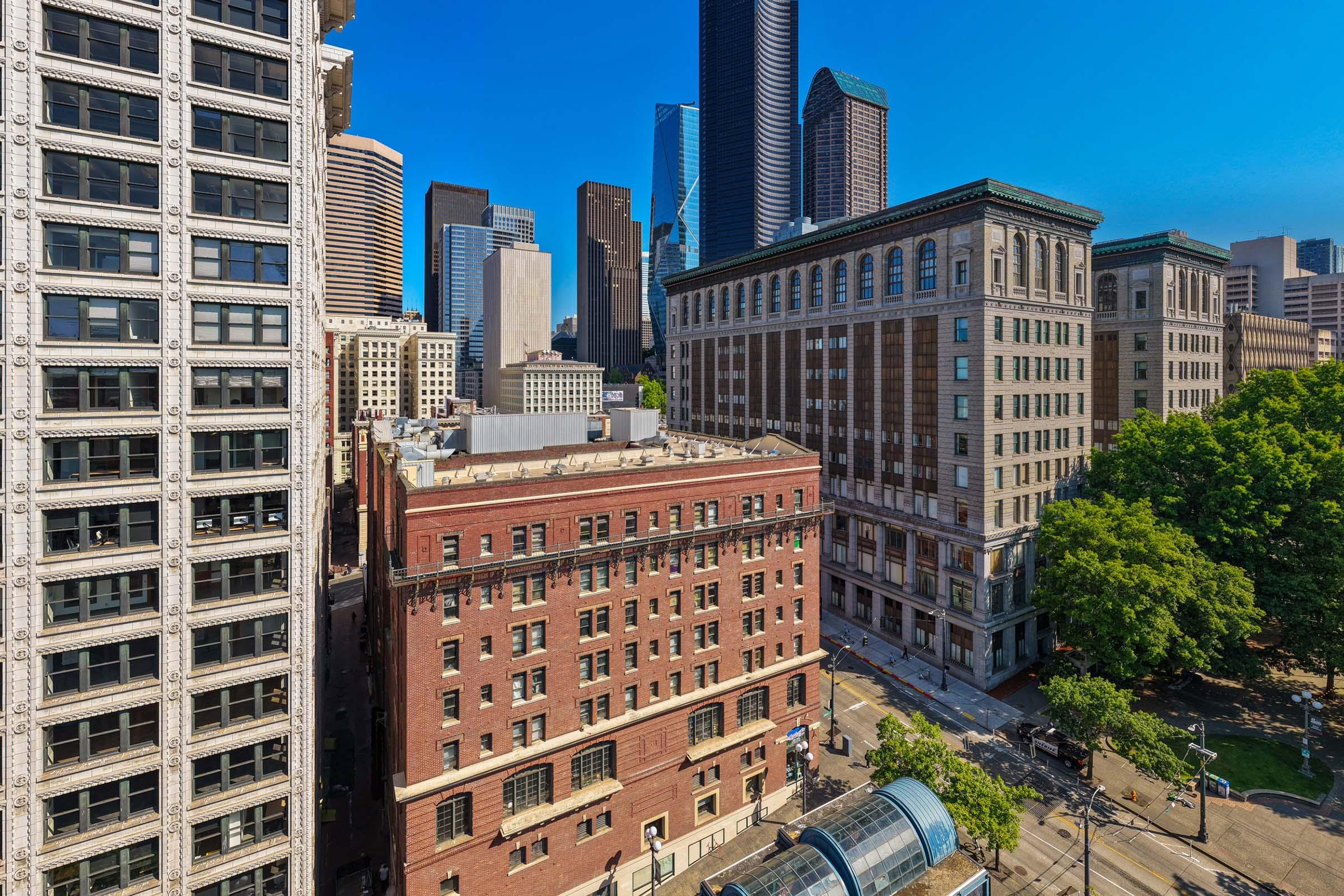 A panoramic view of a cityscape featuring a mix of modern skyscrapers and historic buildings. Notable structures include a brick building with large windows and intricate architectural details, alongside sleek glass towers against a clear blue sky. Trees and a street are visible in the foreground.