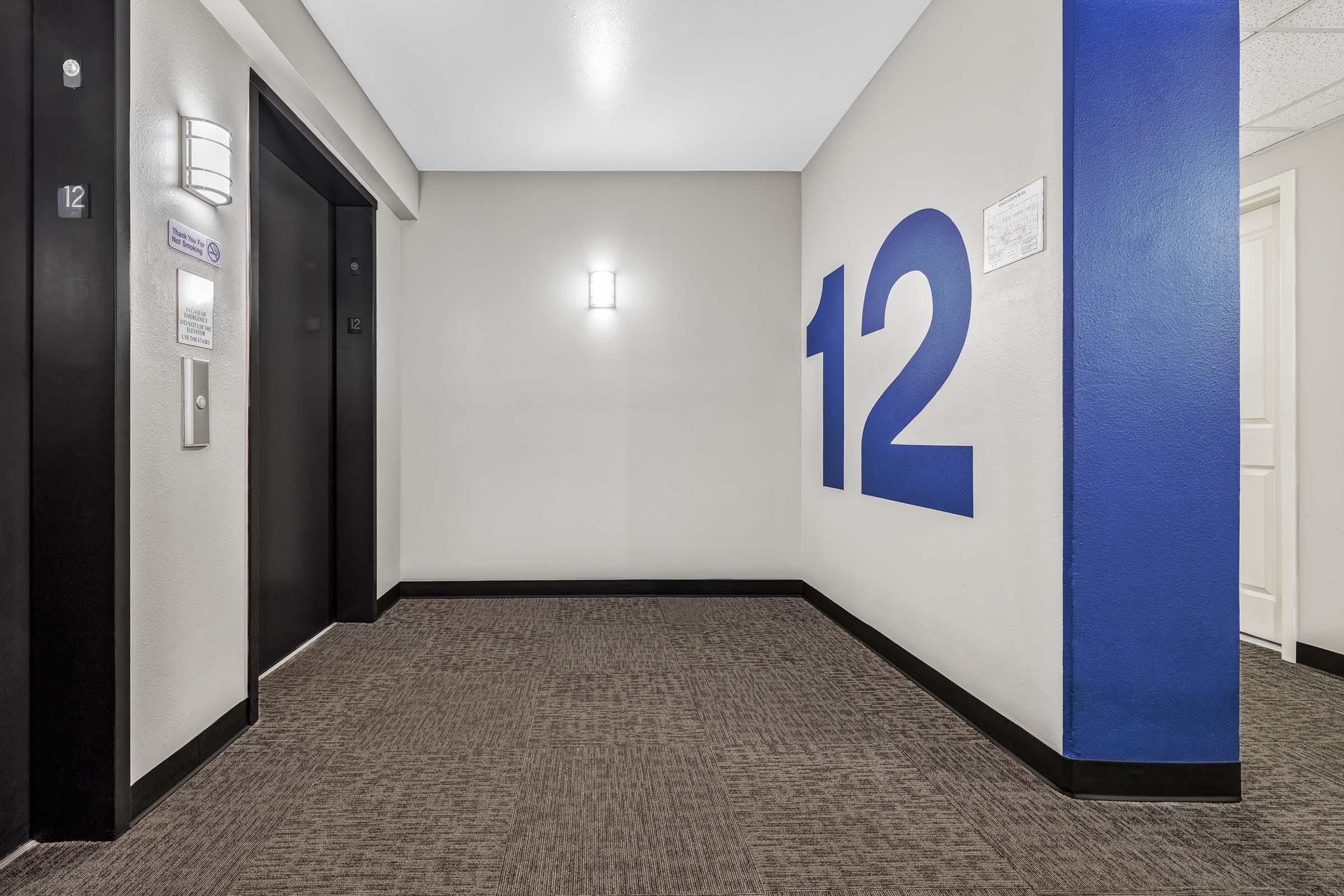 A well-lit hallway featuring elevator doors on the left and a large blue number "12" on the right wall. The flooring is carpeted in a neutral tone, and there is a modern light fixture on the wall. A notice is displayed next to the elevators, enhancing the contemporary design of the corridor.