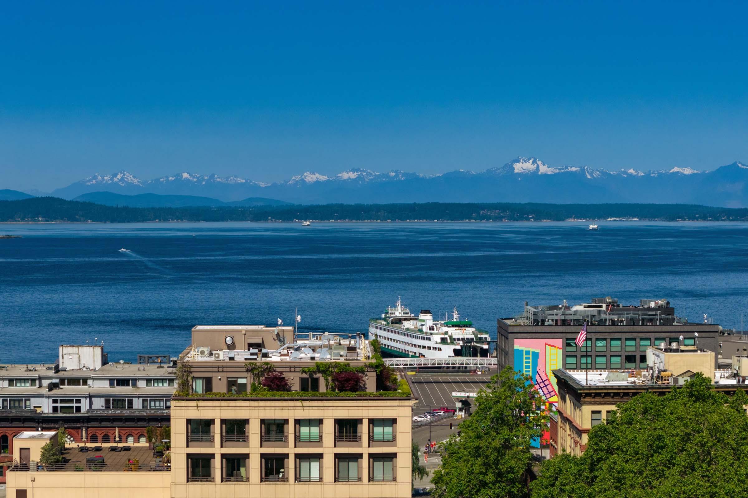 A panoramic view of a waterfront scene showcasing a blue ocean with a ferry in the foreground, surrounded by buildings. In the distance, snow-capped mountains rise against a clear blue sky, adding a scenic backdrop to the bustling urban area.