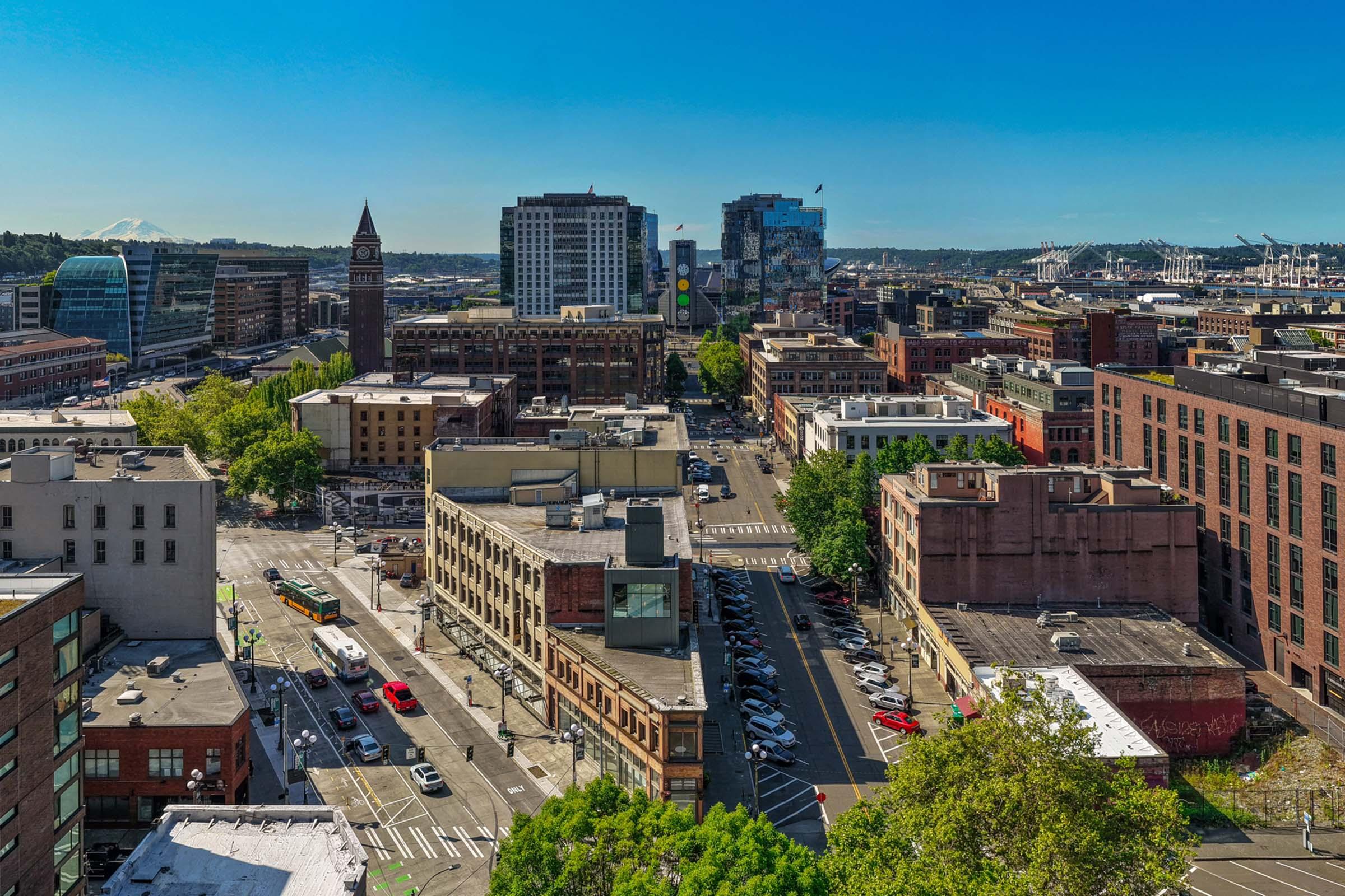 A panoramic view of a bustling cityscape featuring modern skyscrapers and historical buildings. In the foreground, streets are lined with greenery and parked cars, while the background showcases a clear blue sky and distant mountains. The scene highlights urban life and architectural diversity.