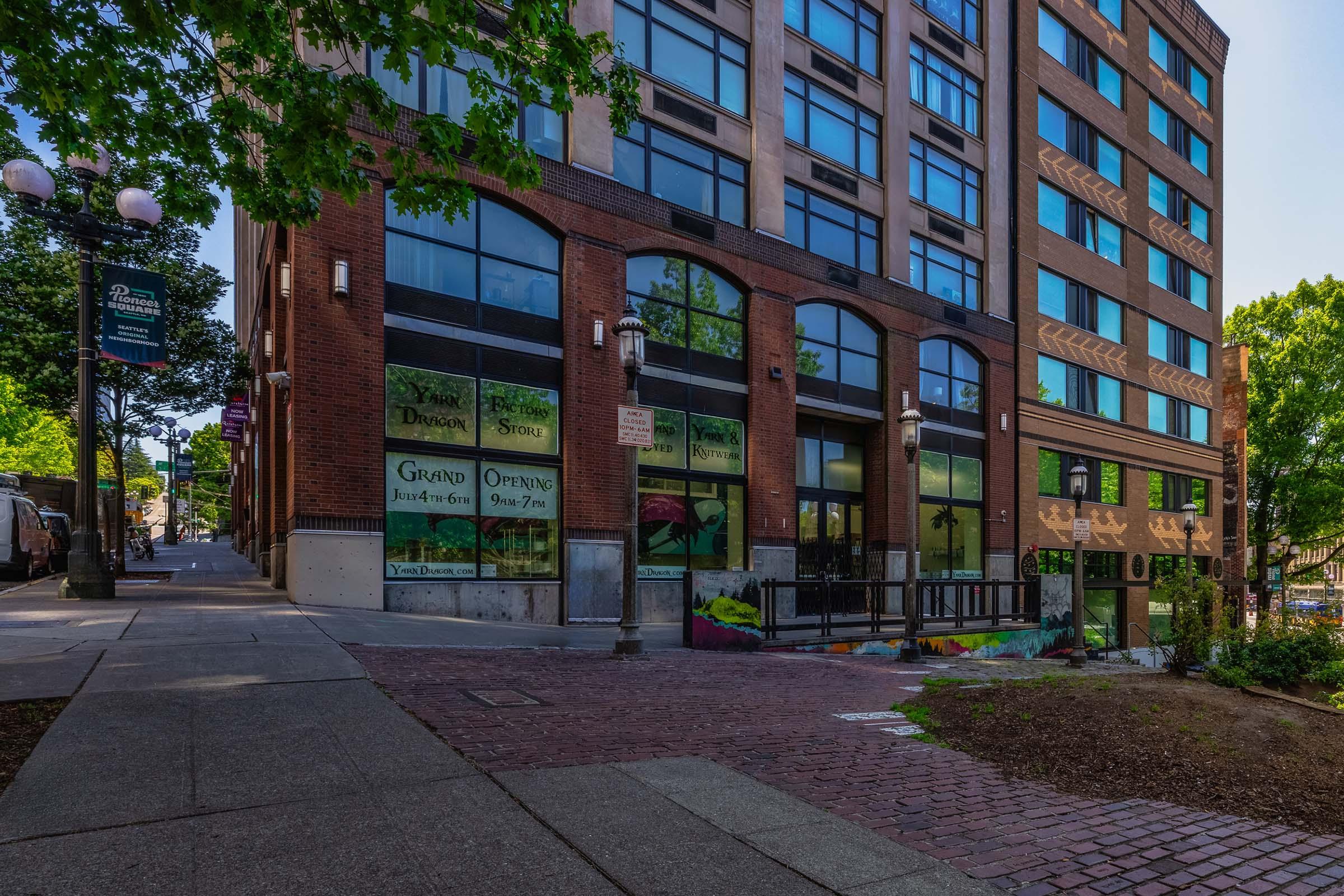 Urban scene featuring a multi-story brick building with large windows. Ground-level storefronts have promotional signage for a grand opening. Surrounding greenery and a brick sidewalk lead up to the building, creating a welcoming city atmosphere.