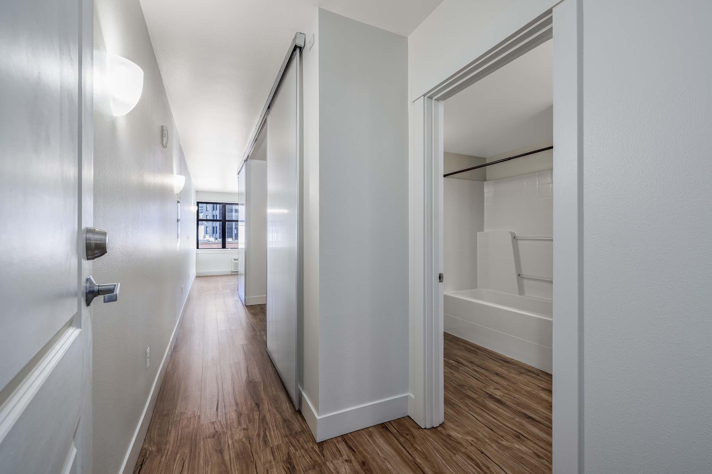 A well-lit interior hallway leading to a bathroom. The walls are painted light gray, and the floor features laminate wood. On the right, a sliding door opens to a bathroom with a bathtub and shower. Natural light enters from a window at the end of the hallway, enhancing the spacious feel.