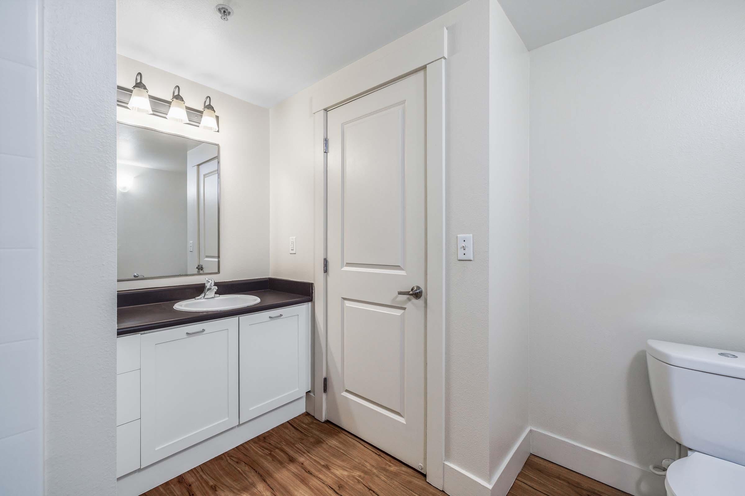 A clean and modern bathroom featuring a white vanity with a sink and mirror, overhead lighting, a closed toilet, and light wood flooring. The walls are painted in a neutral color, and there is a door leading out of the bathroom.